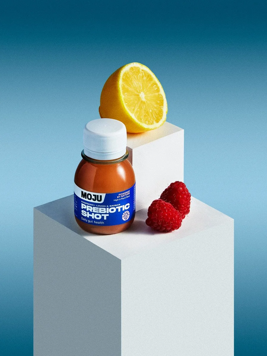 MOJU Prebiotic Shot bottle on white pedestals with a fresh lemon half and two red raspberries for gut health.