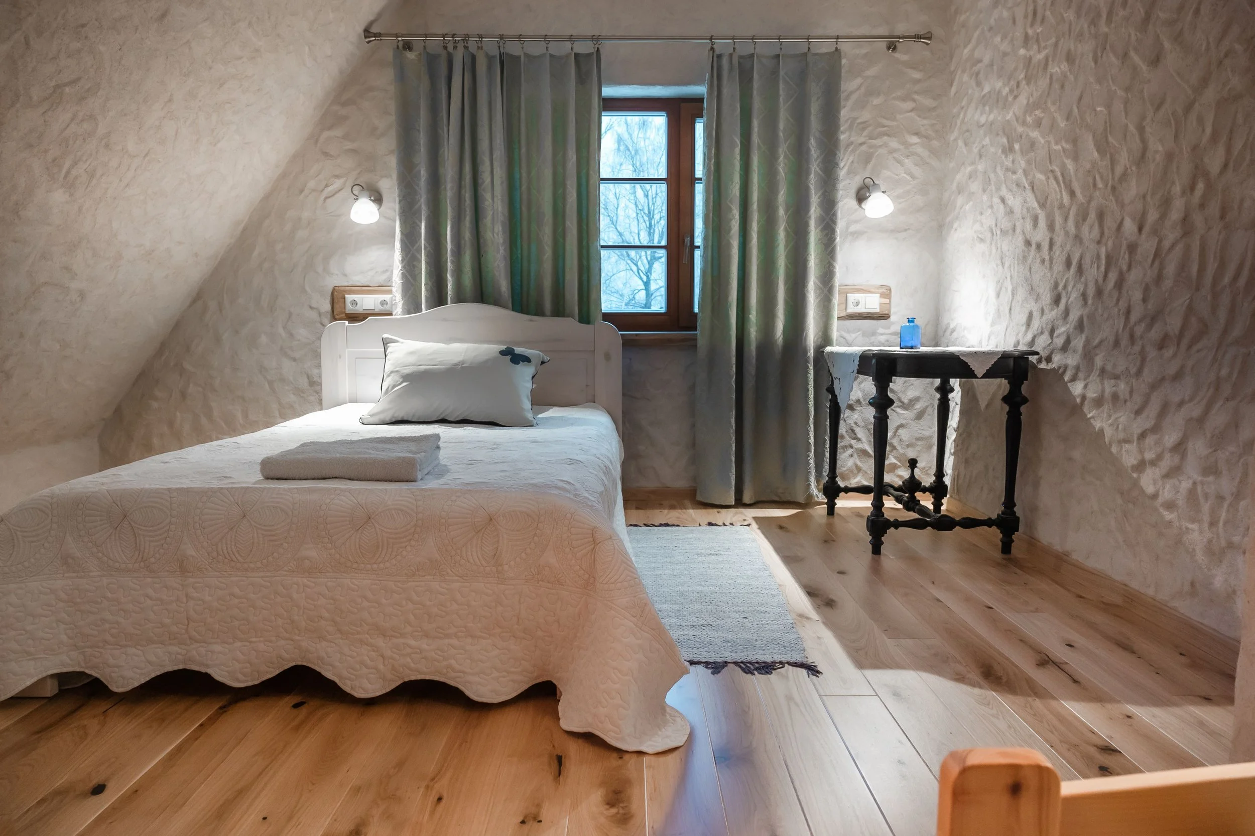 A cozy bedroom with textured white walls, a wooden-framed window with green patterned curtains, and a bed with white bedding and a pillow with a butterfly design. A small black table with a blue water bottle is beside the window, and two wall-mounted