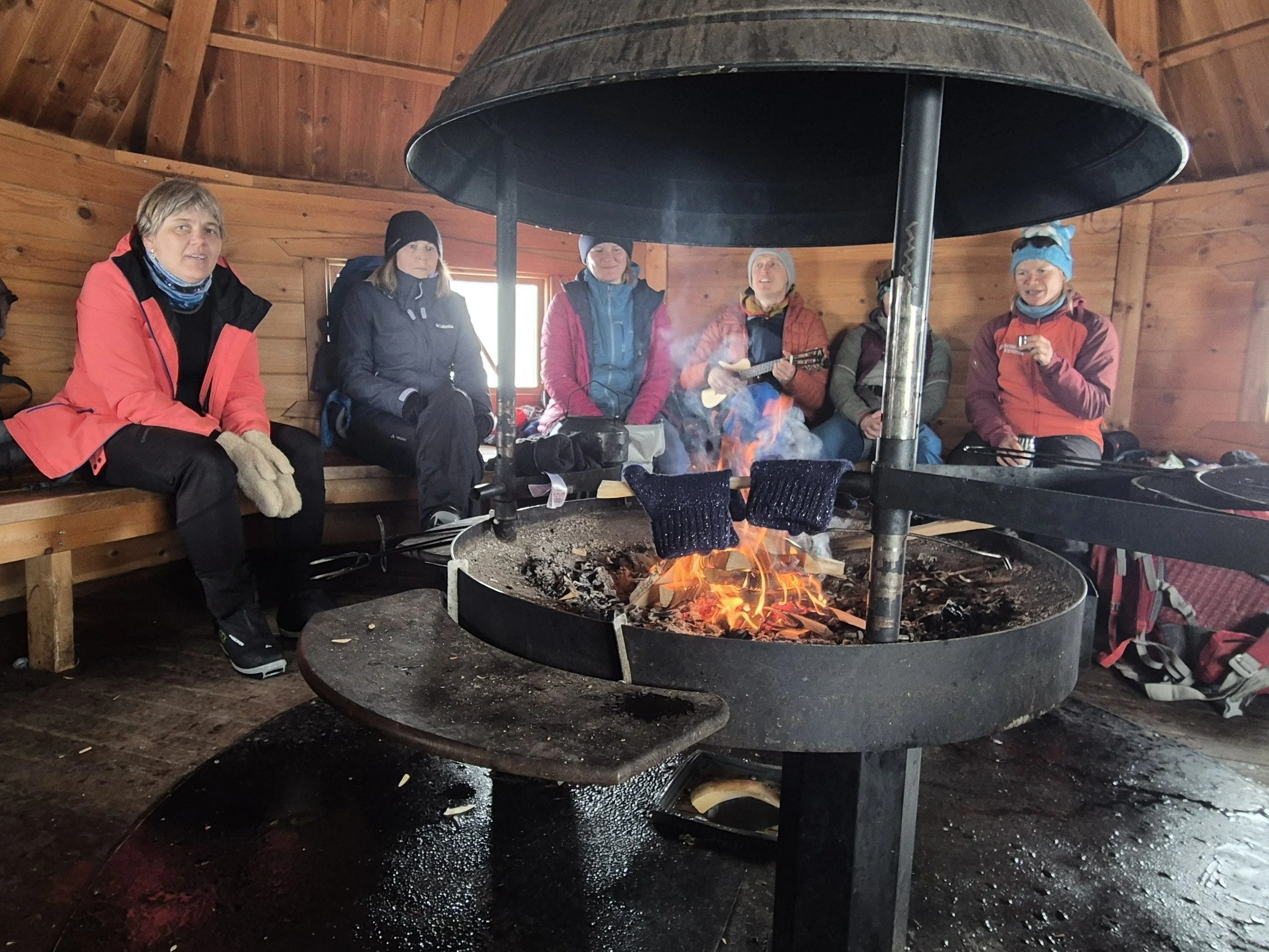 Group of six women wearing winter clothing sitting around a fire inside a wooden shelter, some holding musical instruments and drinking.