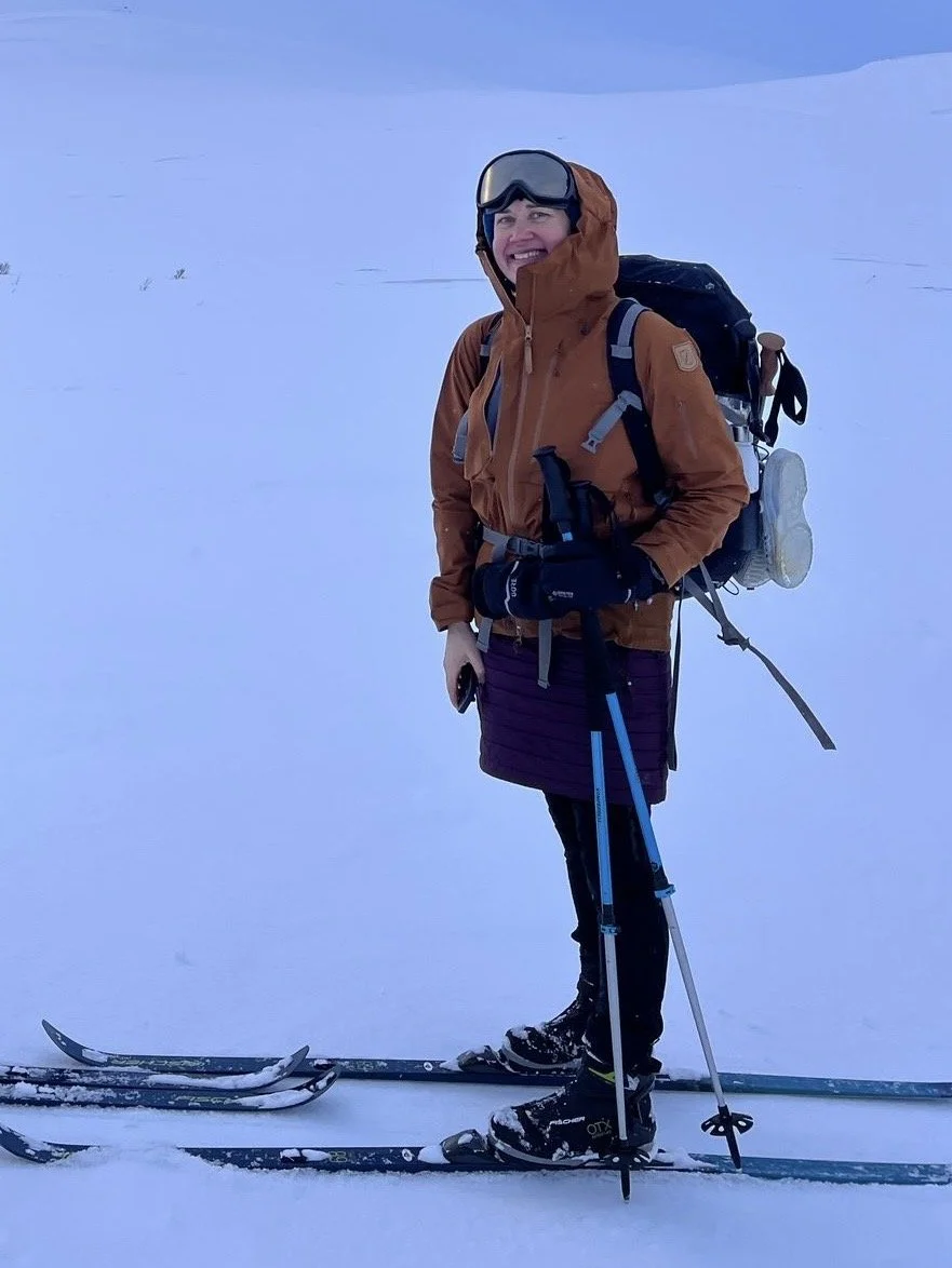 A group of people hiking through snowy woods, with a woman wearing a bright green jacket and orange hat leading the way, using trekking poles.