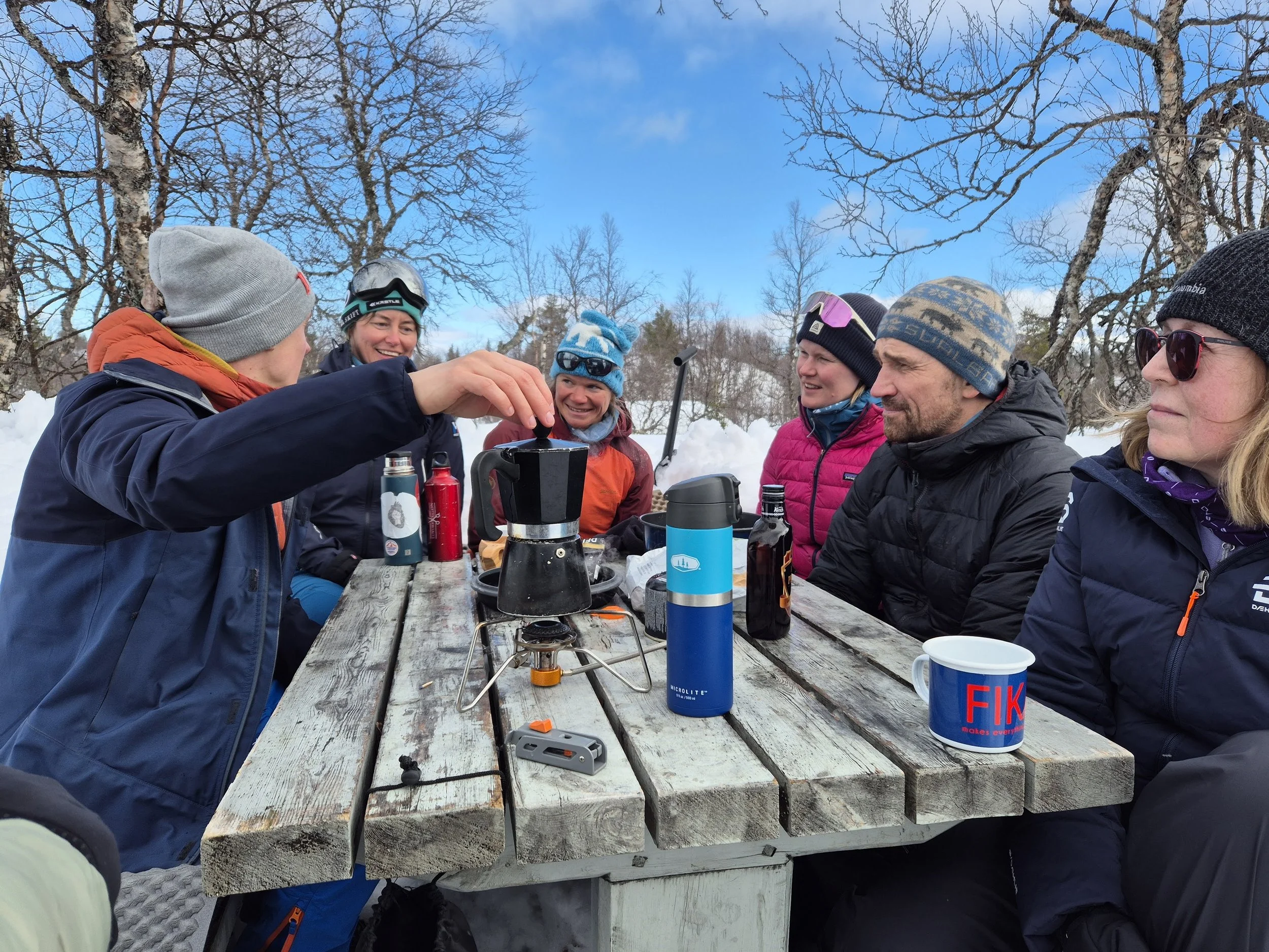 Group of six people enjoying outdoor winter gathering at a wooden picnic table, surrounded by snow and leafless trees, with a clear blue sky overhead.