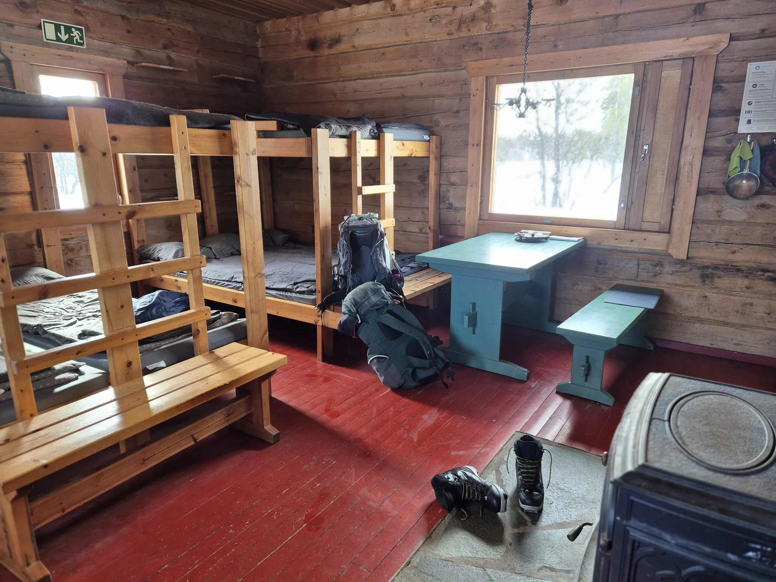 Interior of a wooden cabin room with bunk beds, a blue table, and hiking backpacks, shoes, and gear on the floor near a window and a wood-paneled wall.