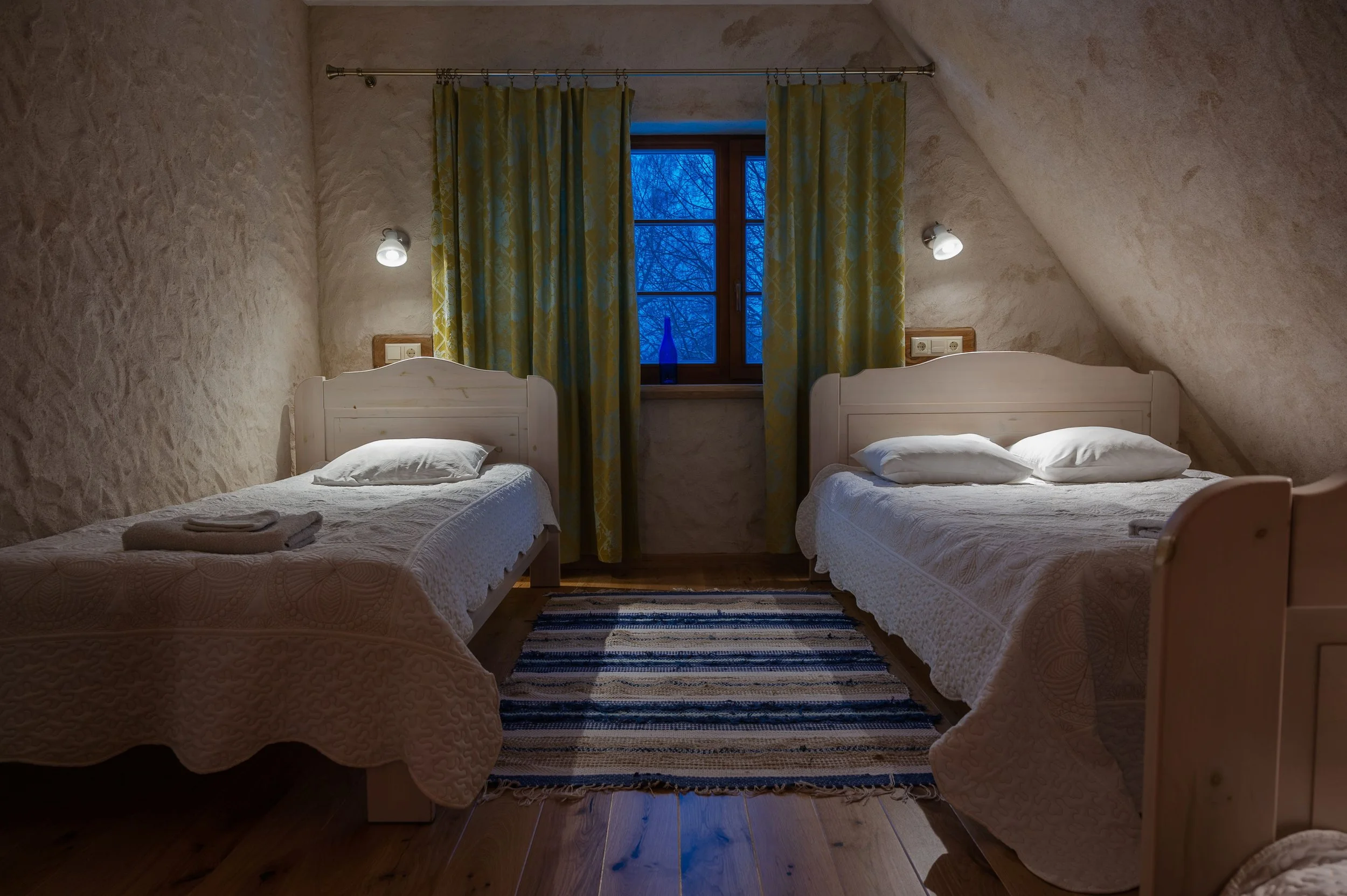 A cozy bedroom with two single beds covered in white bedding, positioned on either side of a window with blue curtains. The room has slanted ceilings with textured beige walls, two wall-mounted lights above each bed, and a dark striped rug on a woode