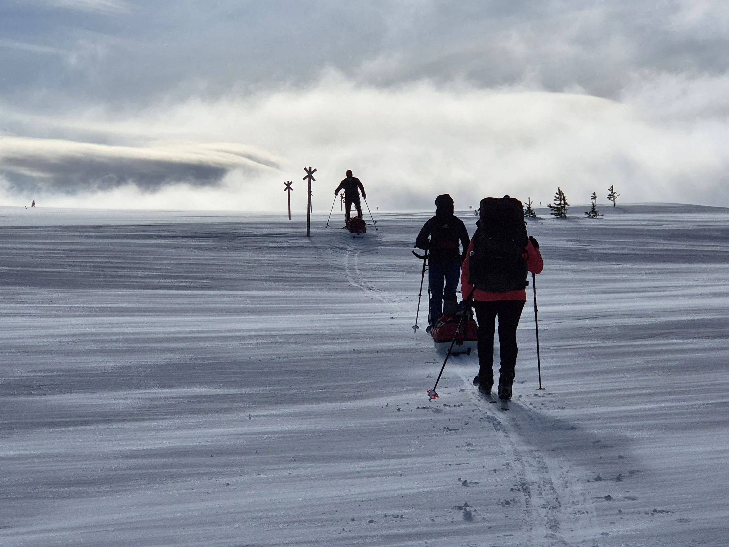 Two hikers walking through snow-covered landscape with snow plows in the distance, under cloudy sky.