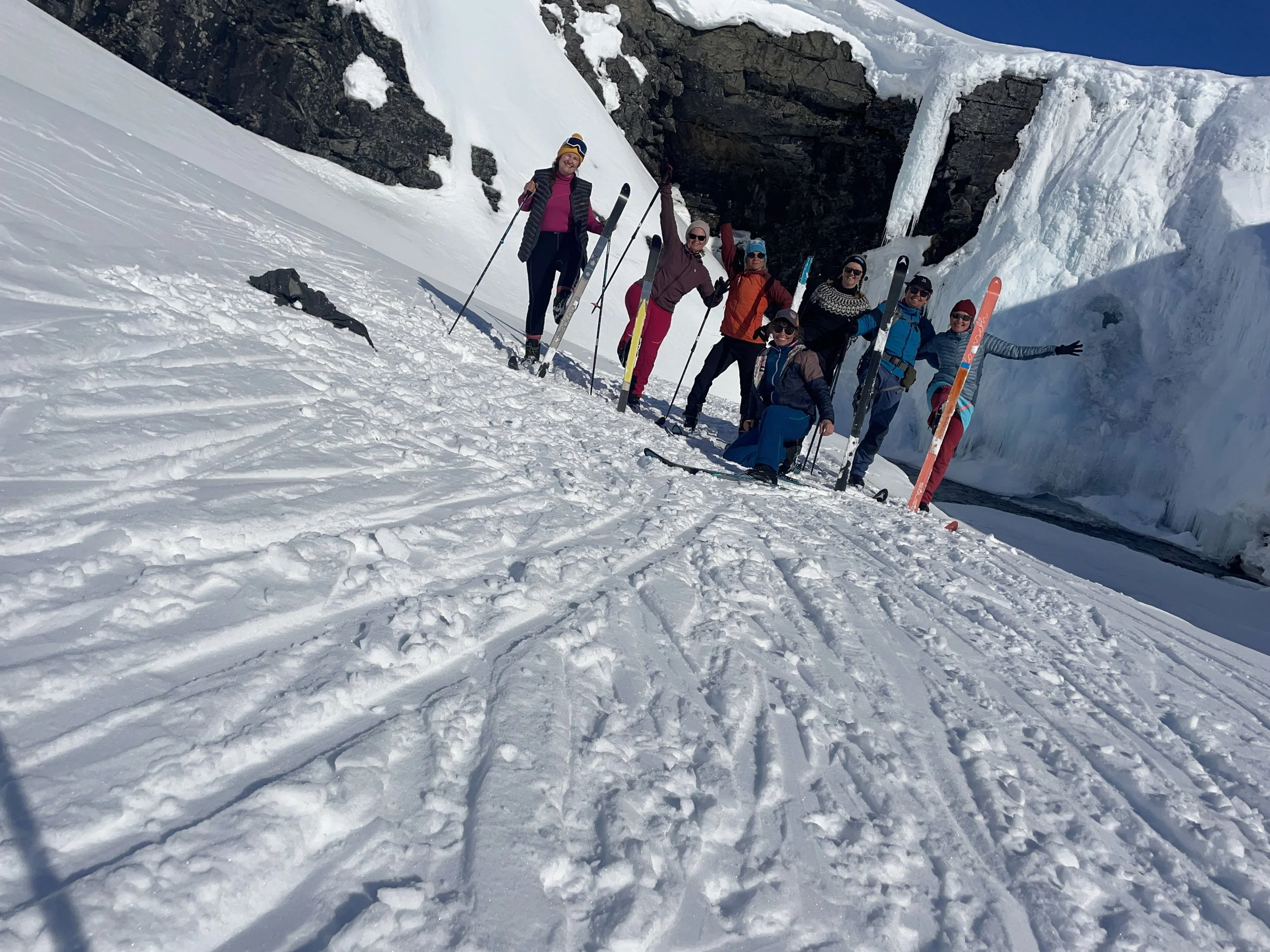 Group of people in winter clothing standing on snow-covered ground with skis and poles, posing for a photo in a snowy mountain landscape.