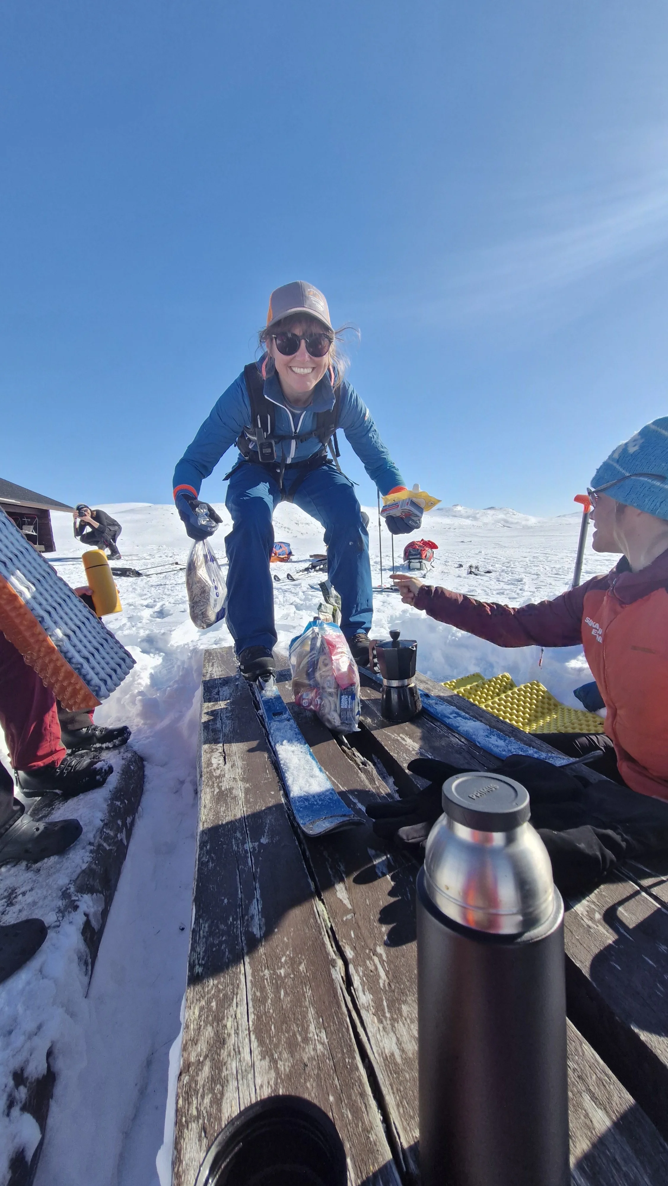 A smiling woman in sunglasses and outdoor winter clothing crouches on skis on a wooden platform in a snowy mountainous area, surrounded by other people and equipment, with a clear blue sky overhead.
