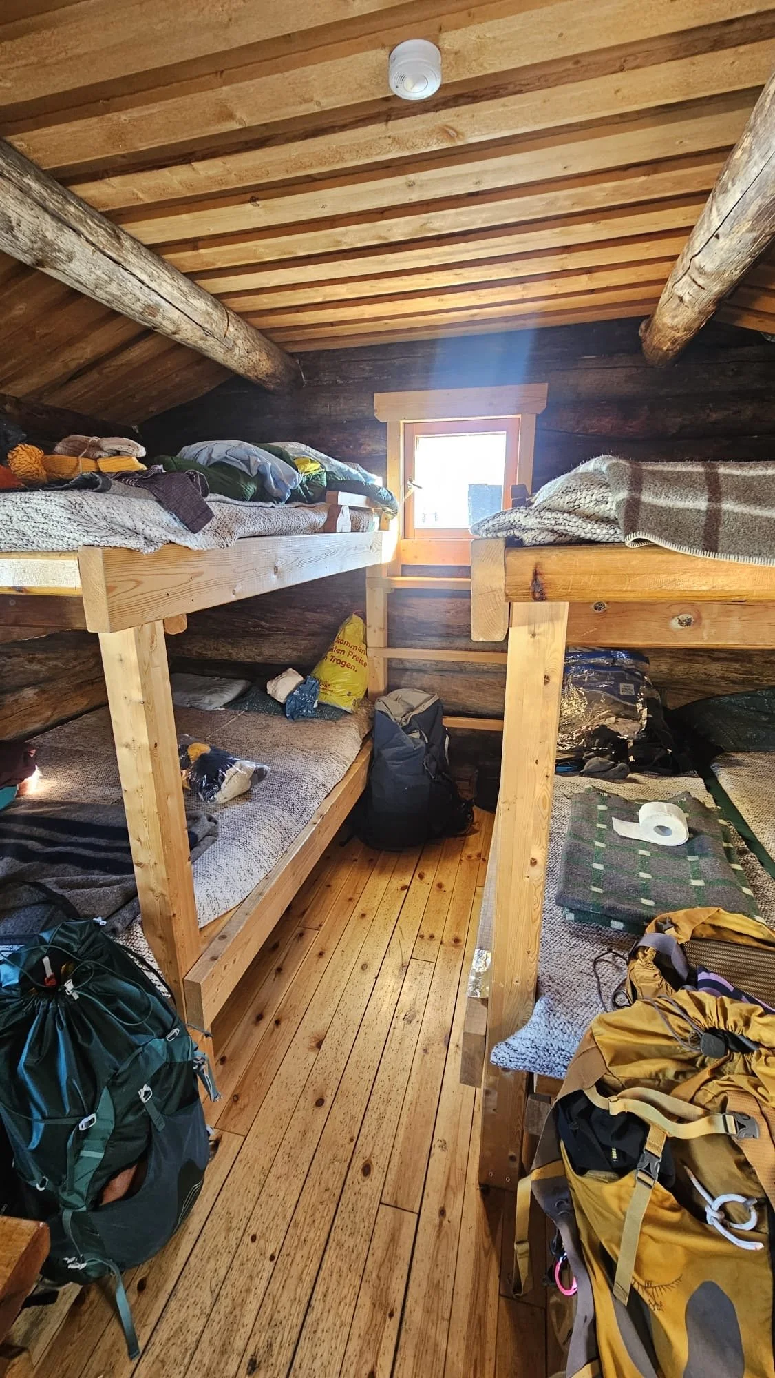 Interior of a rustic wooden cabin with two bunk beds, scattered clothing and gear, and a small window on the back wall.