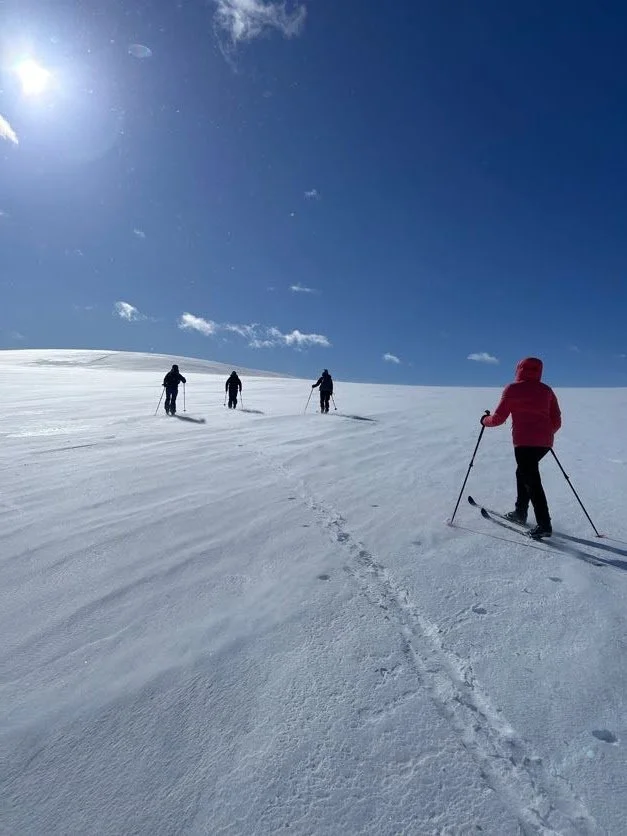 People cross a snowy landscape on skis under a clear blue sky with a few clouds.