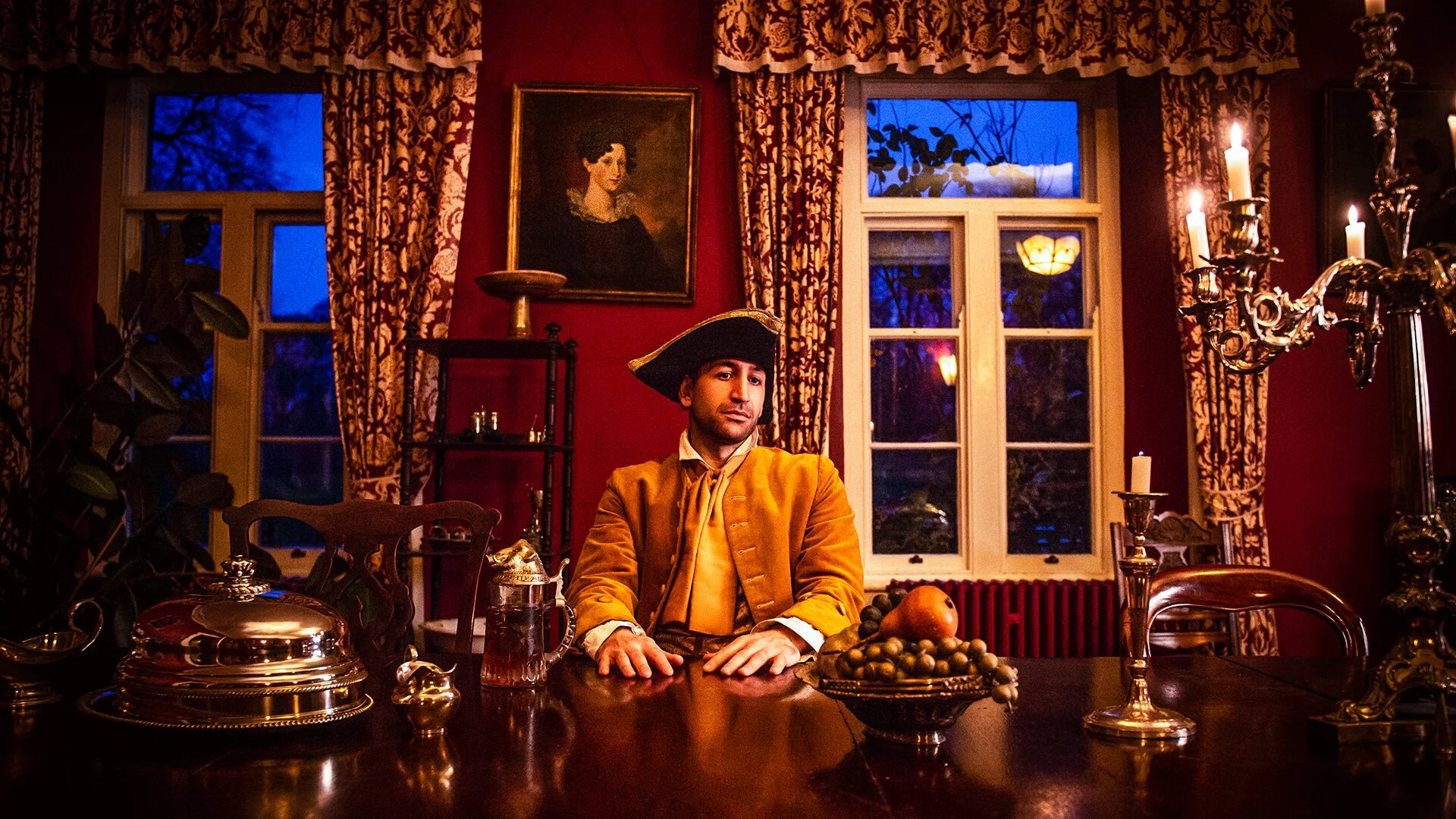 A man dressed as a colonial-era figure, wearing a tricorn hat and a mustard-colored coat, seated at a large wooden table in a richly decorated dining room with red walls, ornate curtains, and a chandelier with lit candles. There are plates of fruit, silverware, and a silver tea pot on the table, and a framed portrait of a woman hangs on the wall behind him.