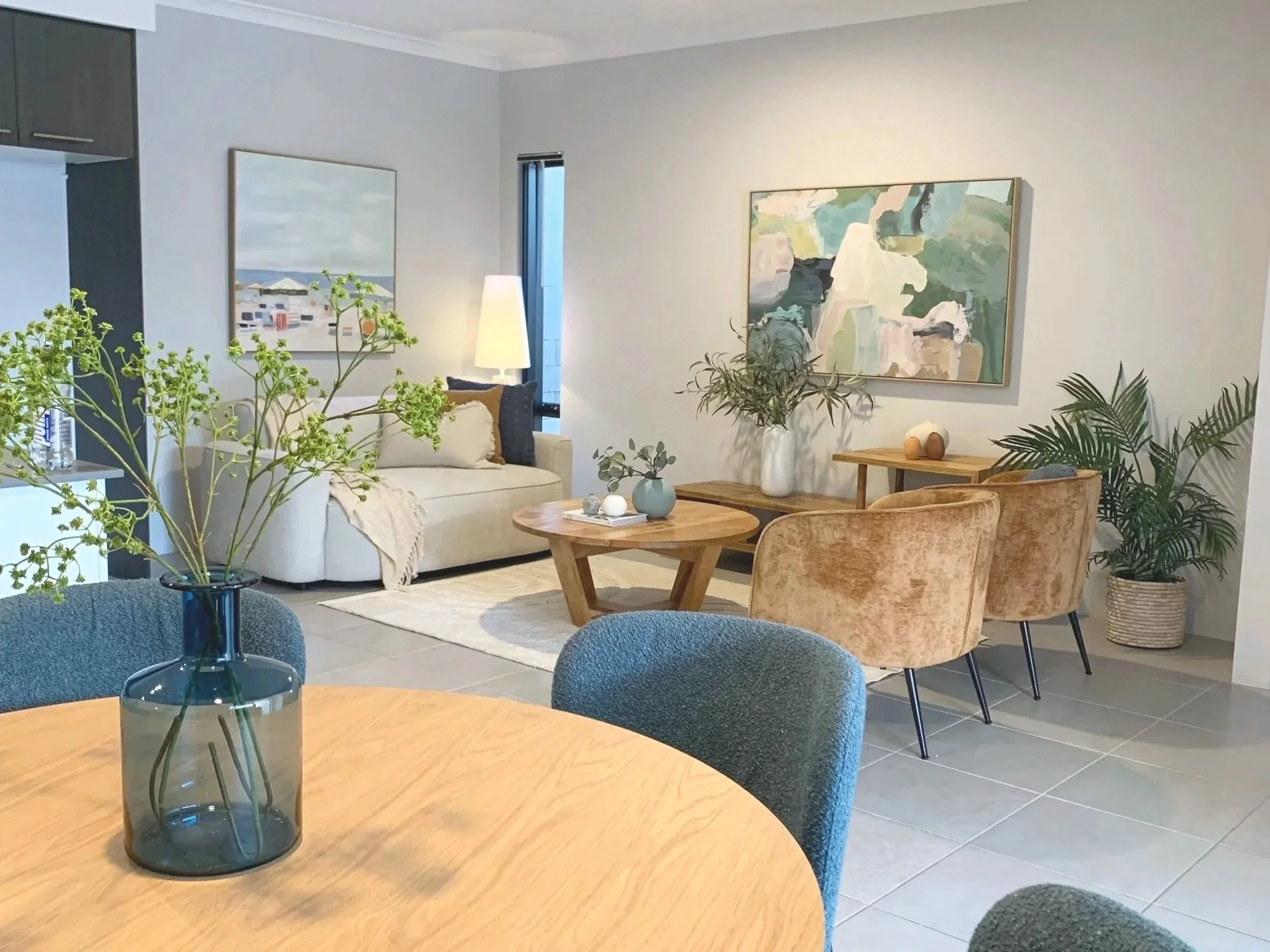 Living room with a round wooden dining table and blue chairs in the foreground, a beige couch with pillows and a throw blanket, a wooden coffee table with vases, and a wooden sideboard with decorative items. Artwork and plants adorn the walls and cor