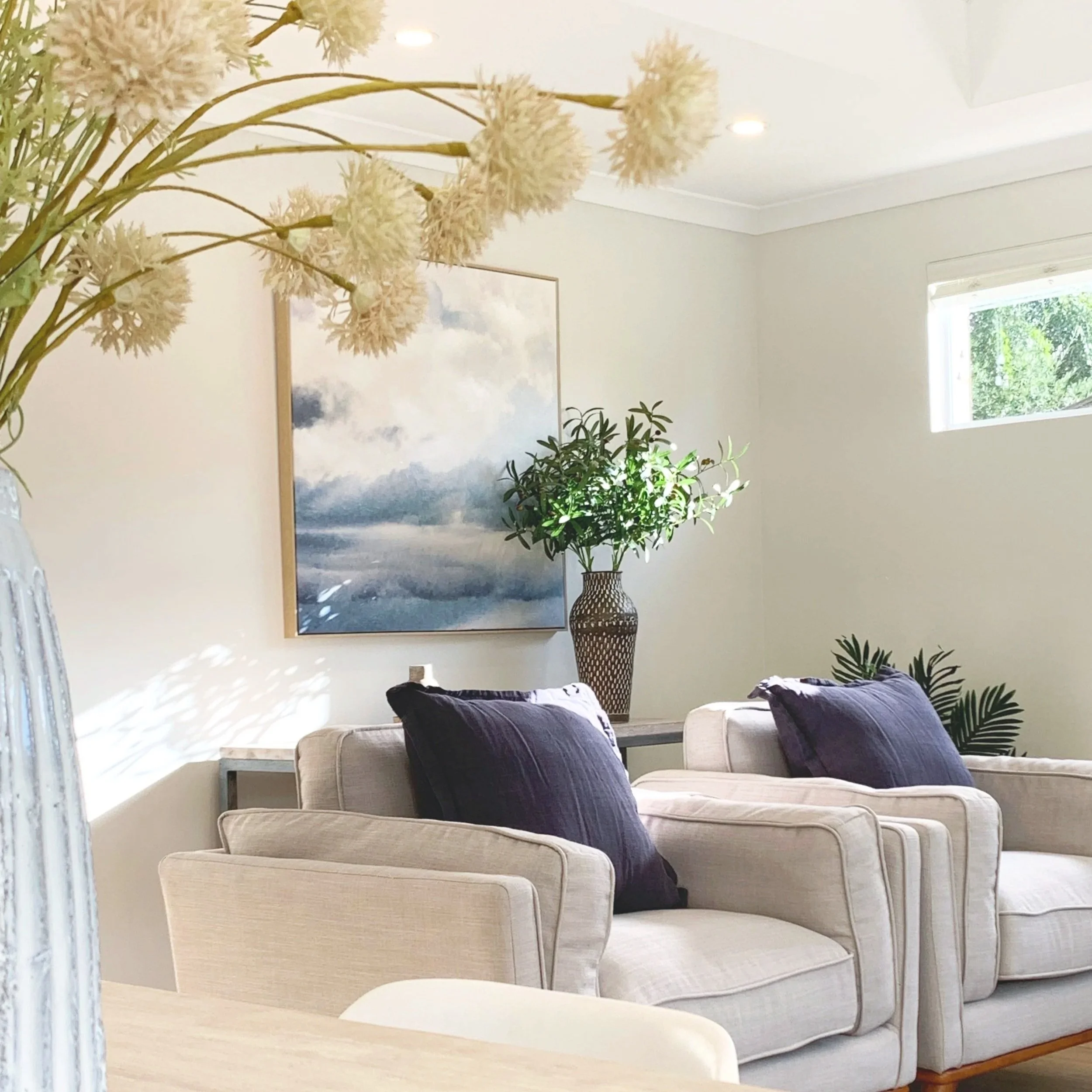 A living room with beige sofas, dark purple cushions, a large green potted plant, a wall art of clouds, and a small window with greenery outside.