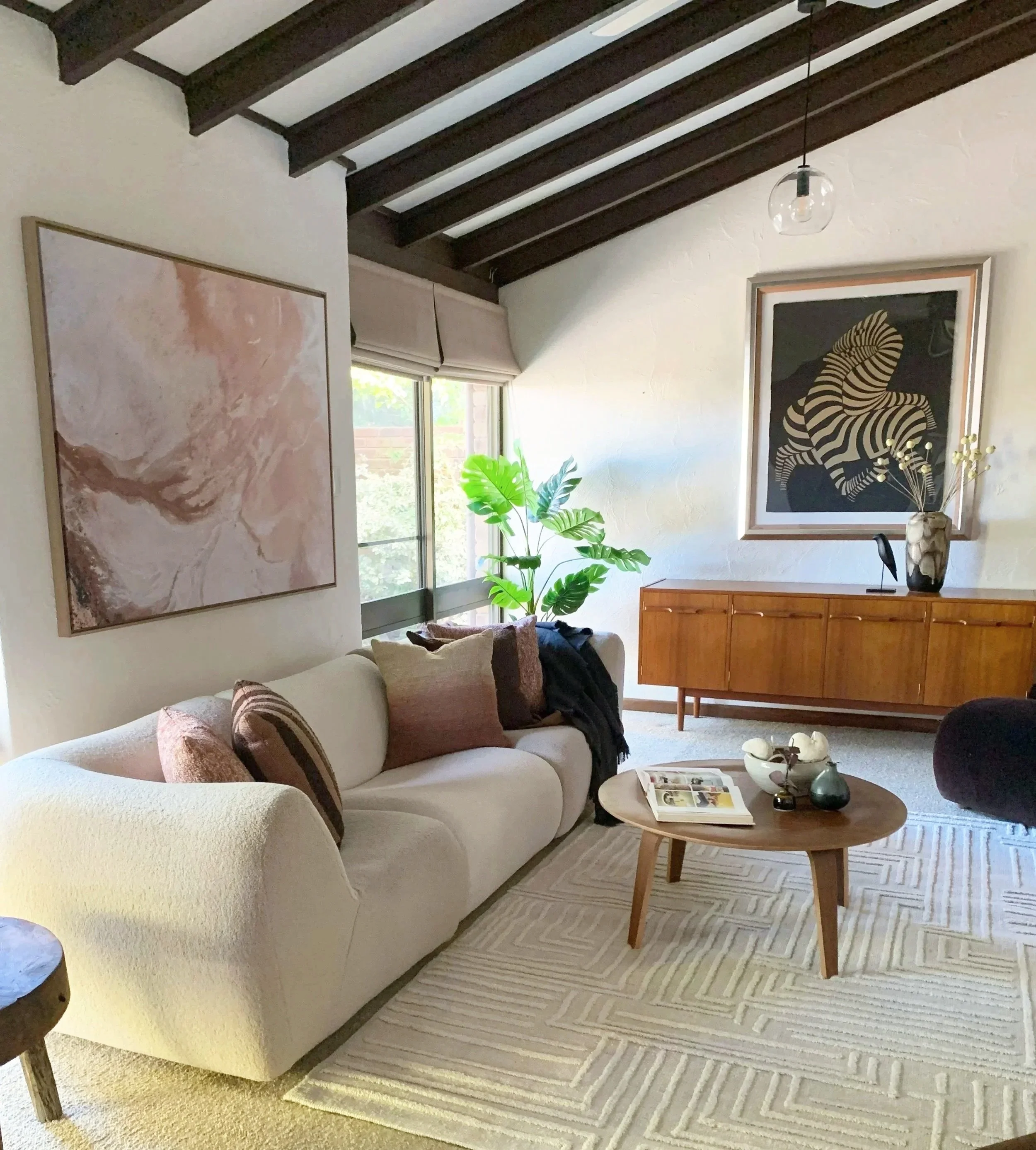 Living room with white sofa, decorative pillows, wooden coffee table with magazines and vases, large window with greenery outside, wooden sideboard, framed artwork on the walls, indoor plant, and a minimalist ceiling light