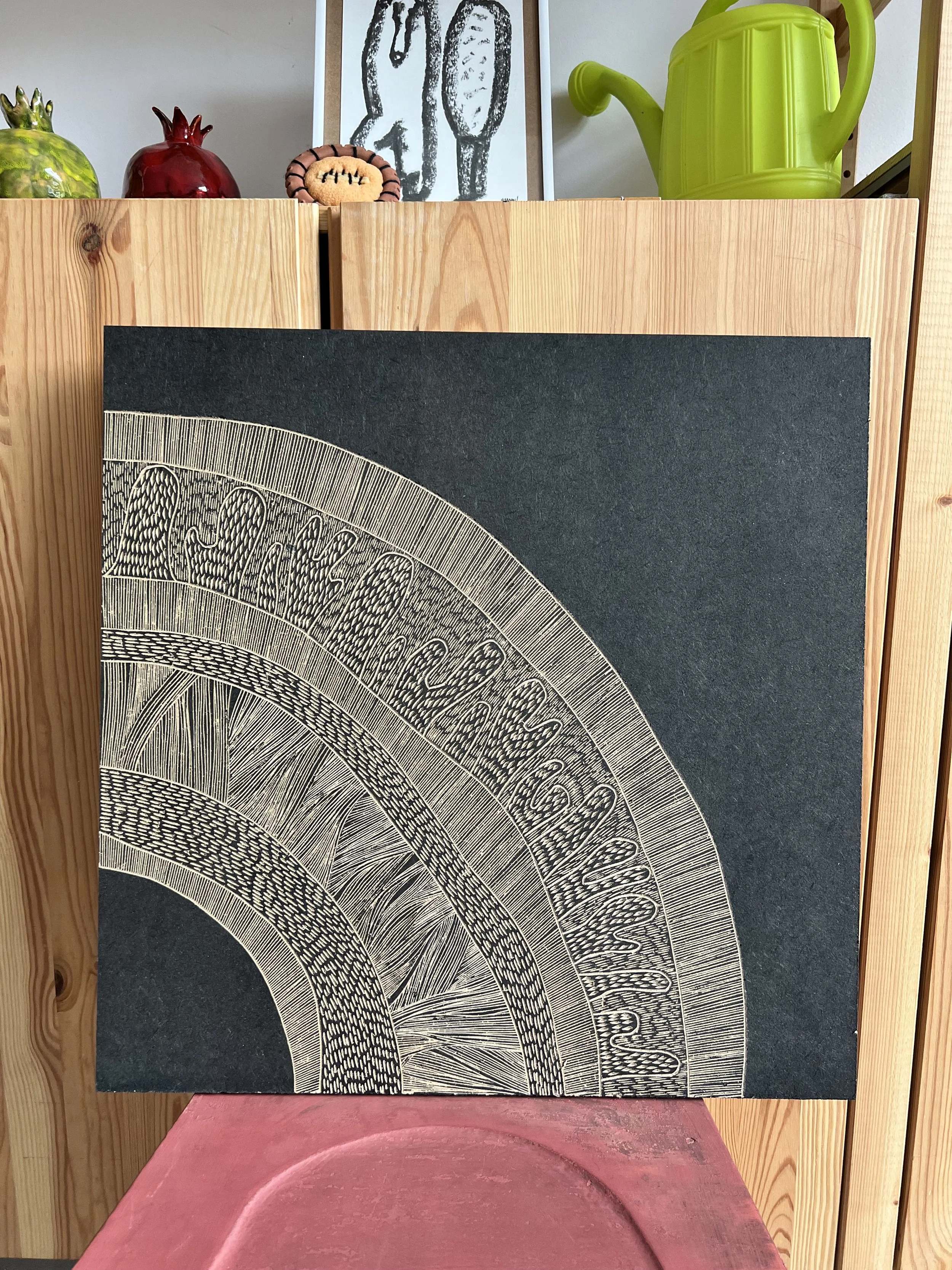 Abstract geometric artwork with intricate line patterns on black canvas, displayed against a wooden backdrop, with decorative objects and a green watering can on top.