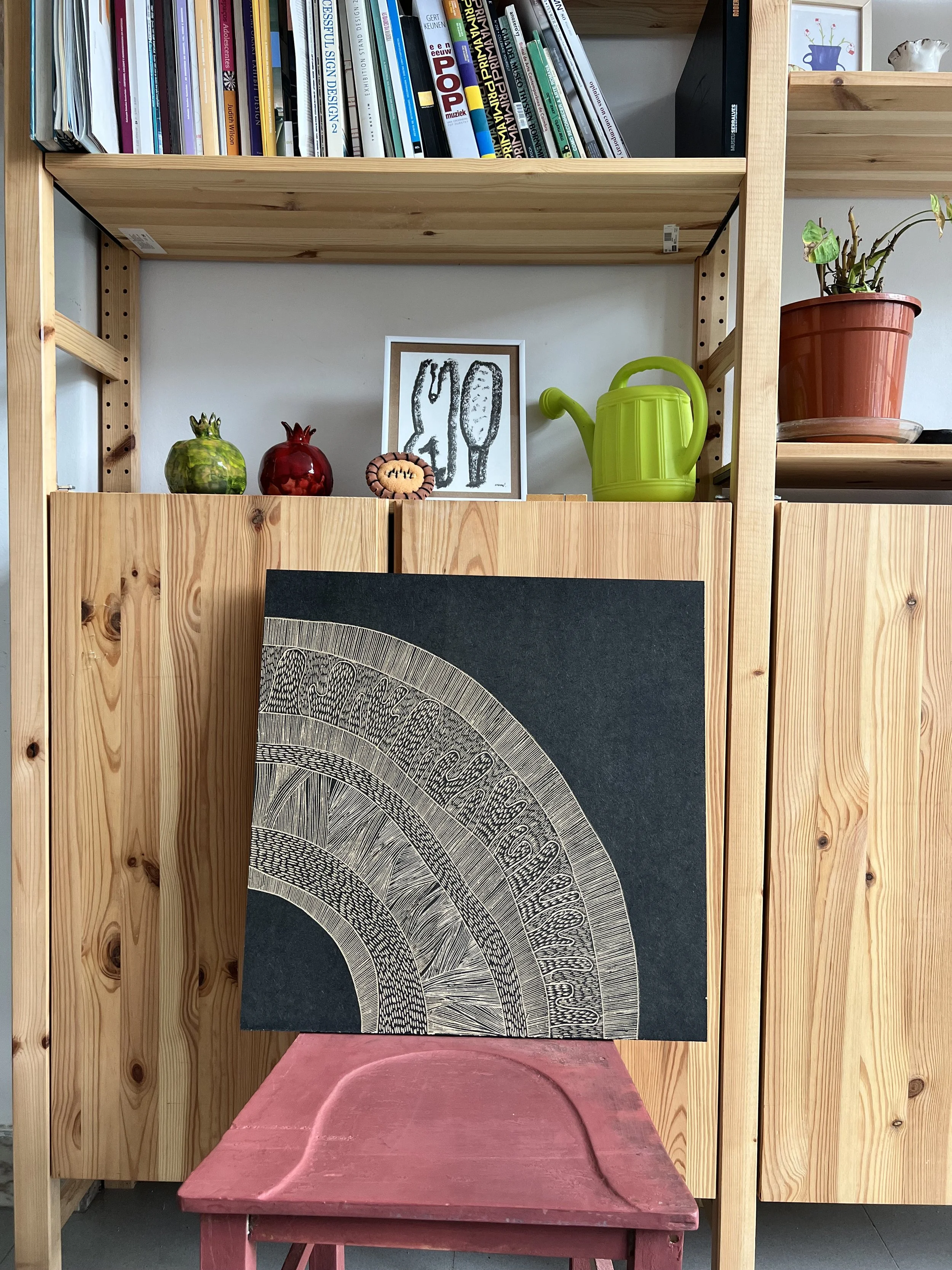Interior scene with a wooden shelf filled with books, decorative items including colorful pomegranate figurines, a small cactus drawing, a green watering can, and a potted plant. A detailed black and beige artwork is resting on a red stool in the foreground.
