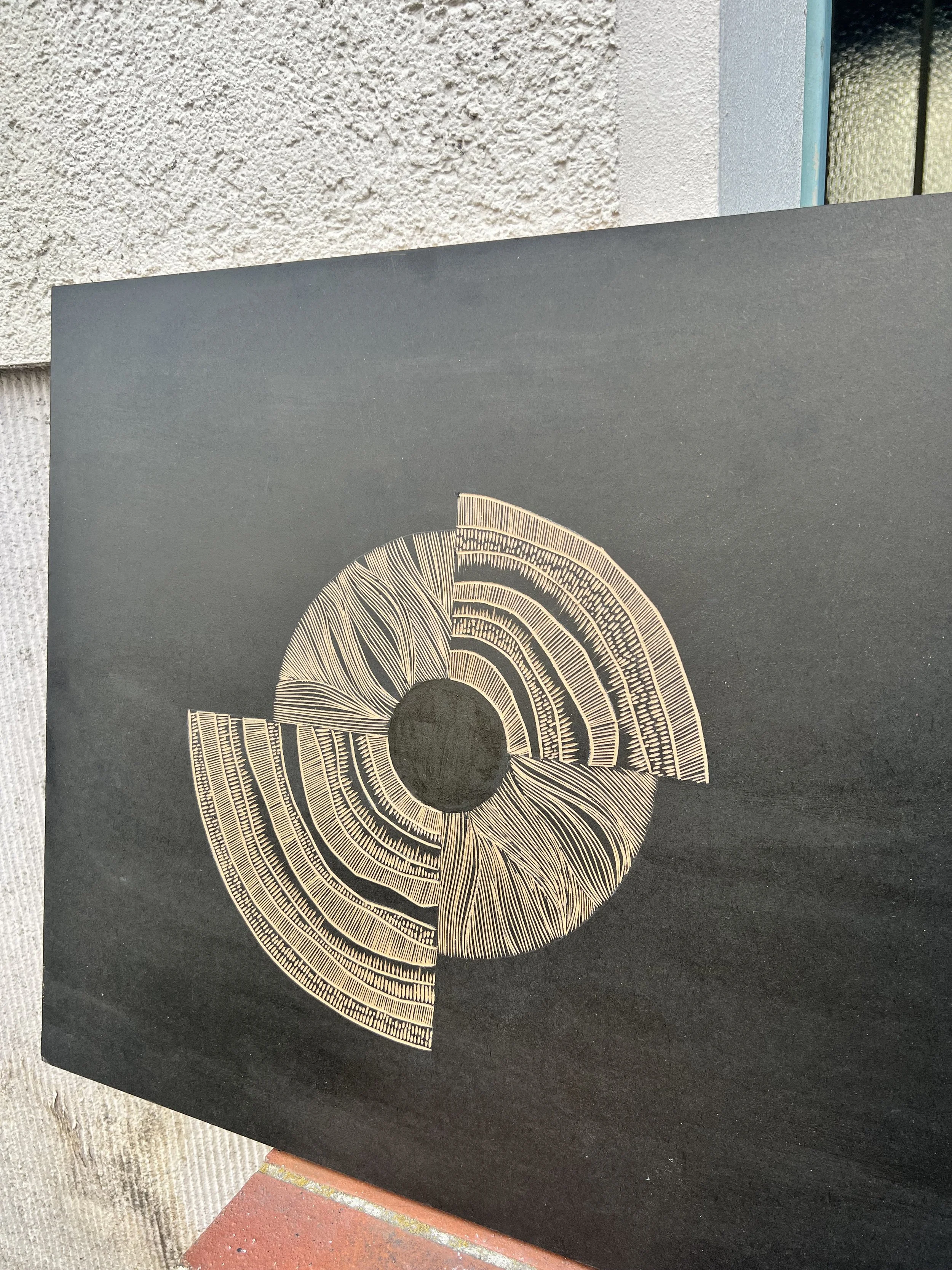 Abstract art on a black canvas featuring a circular design with segmented elements and intricate patterns in a contrasting color. The artwork has a modern and geometric aesthetic, displayed vertically outdoors against a textured white wall.