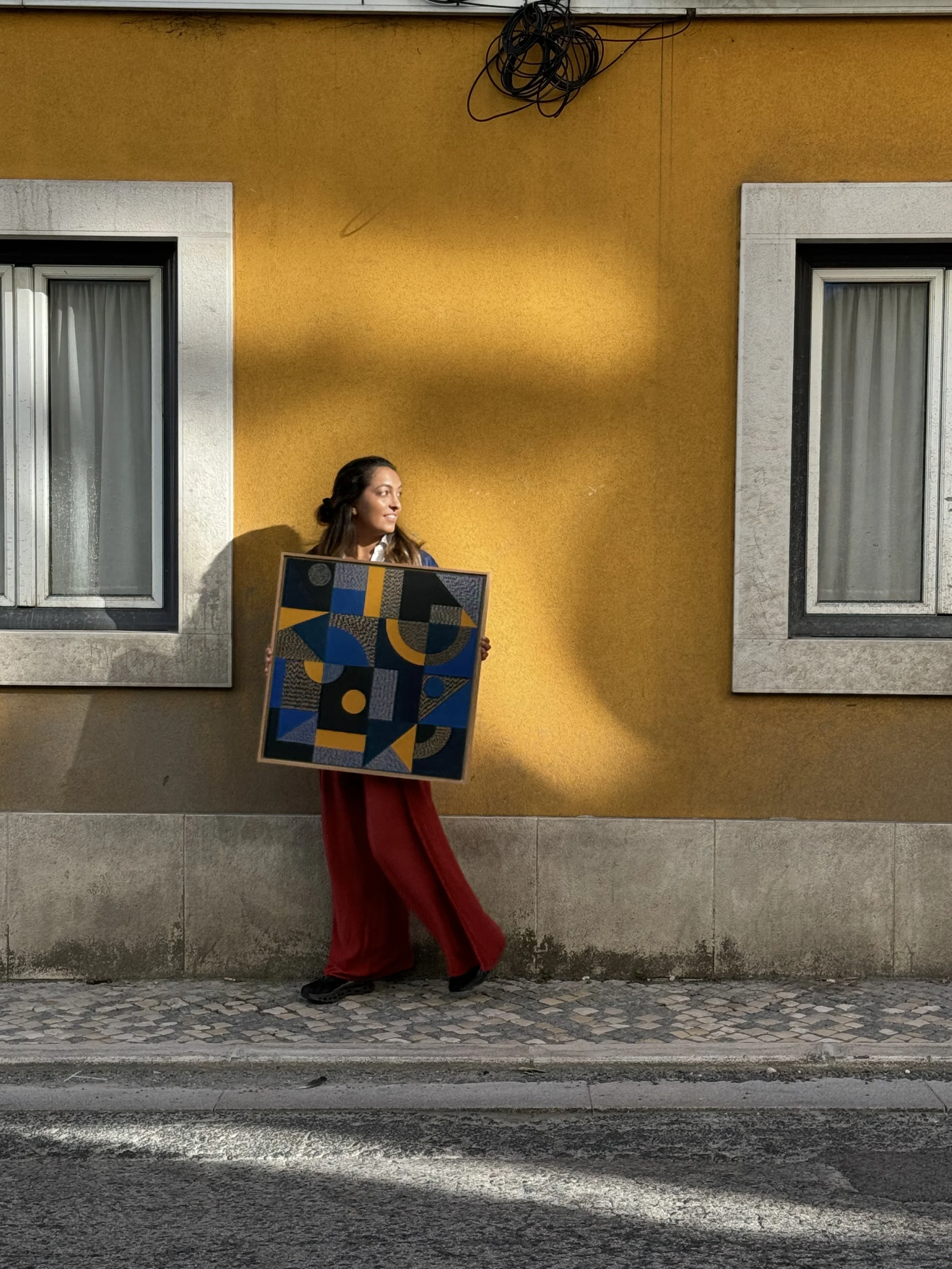 Person holding an abstract painting with blue and yellow geometric shapes, standing in front of a yellow wall between two windows.