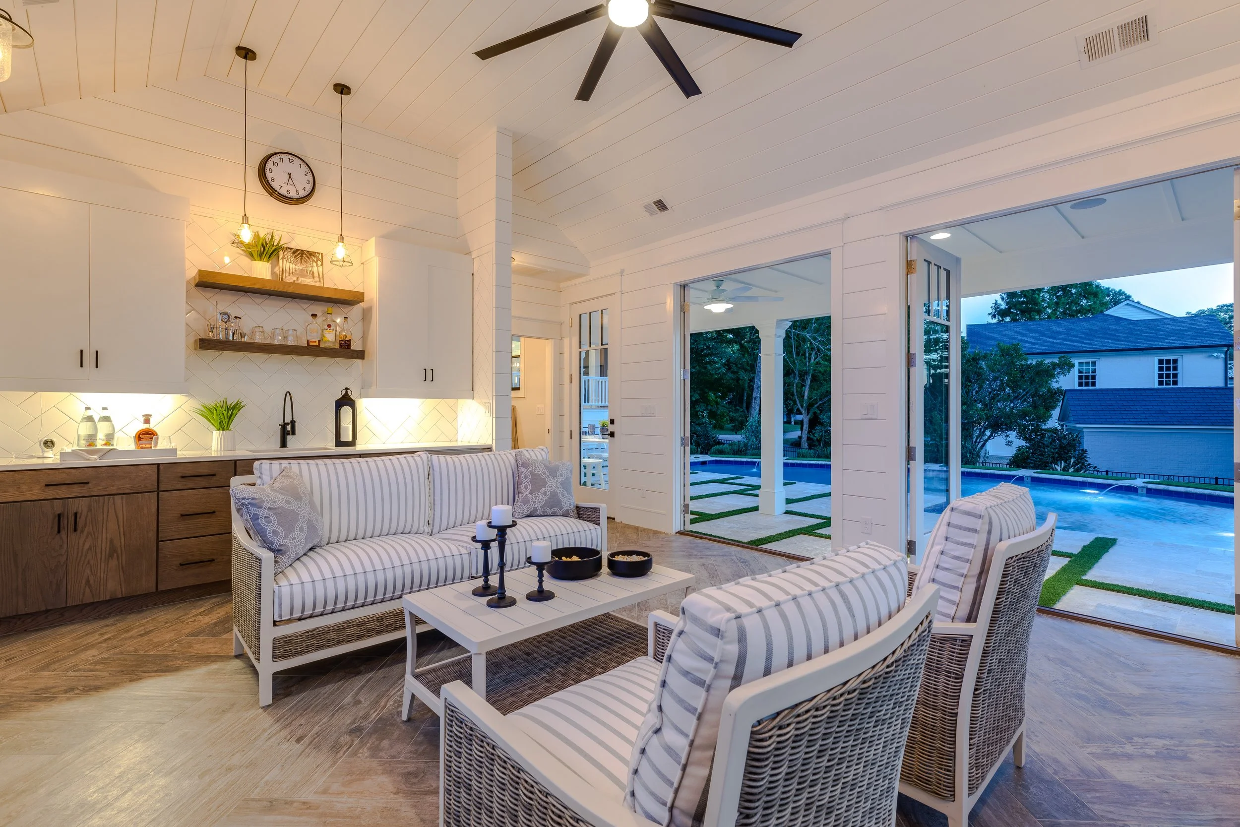 Living room with white shiplap walls, wooden flooring, and a ceiling fan. Open to a patio and backyard with a pool. Seating includes wicker chairs and a sofa with striped cushions. Decor includes candles, black candle holders, potted plants, and shelves with glasses and bottles. Large sliding glass doors open to the outdoor space.