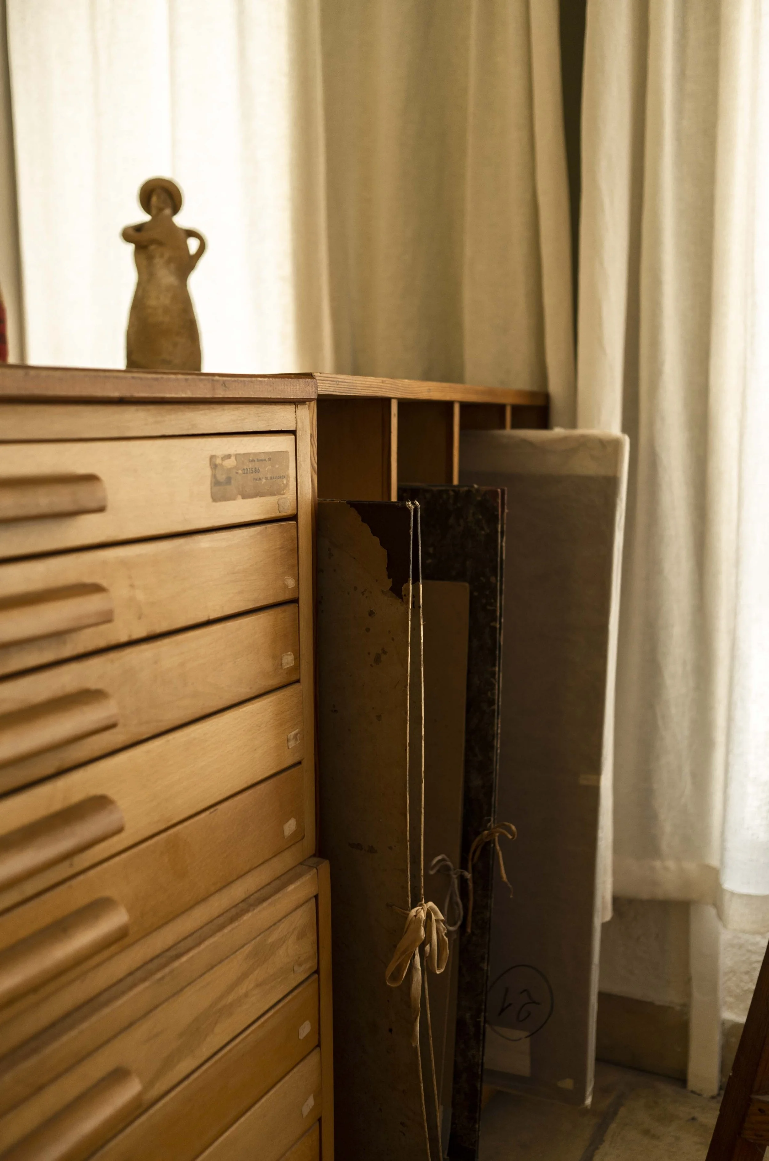A wooden desk with drawers and a small wooden sculpture of a person on top, facing a window with curtains.
