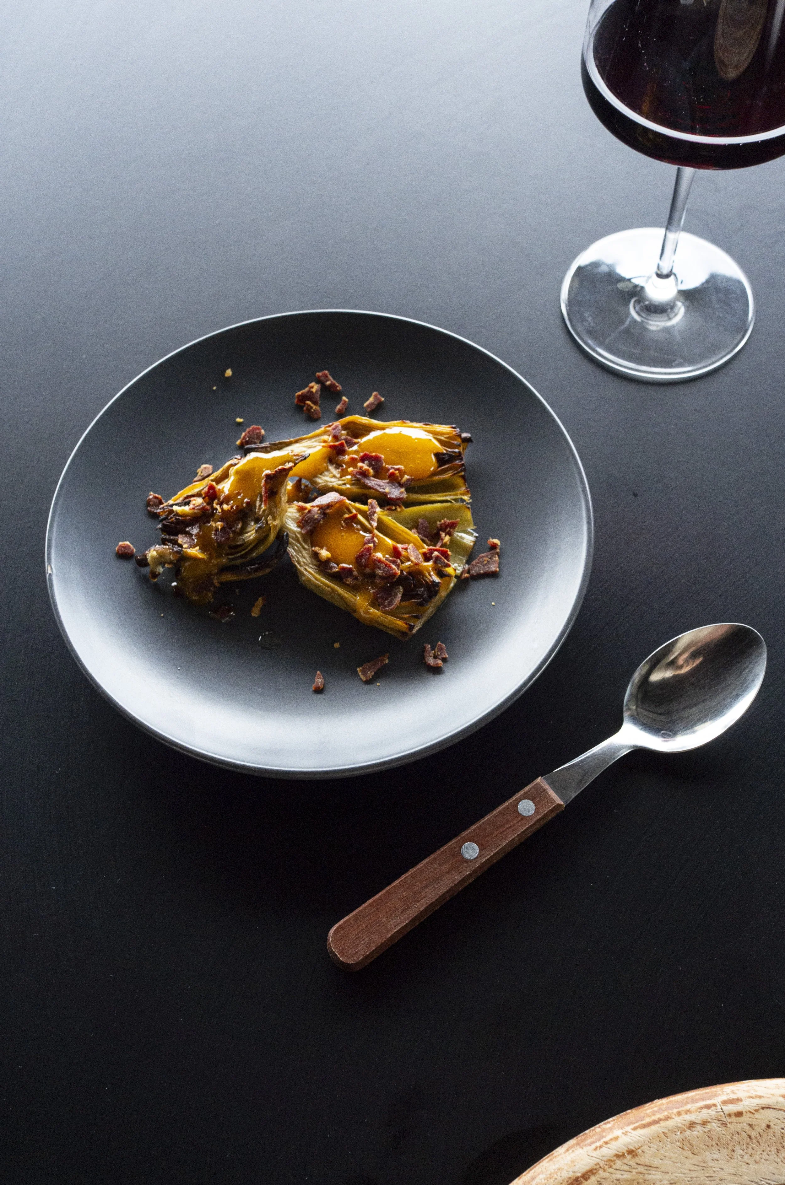 A plate with roasted artichoke halves topped with bacon bits and sauce, a glass of red wine, a spoon with a wooden handle on a dark table.
