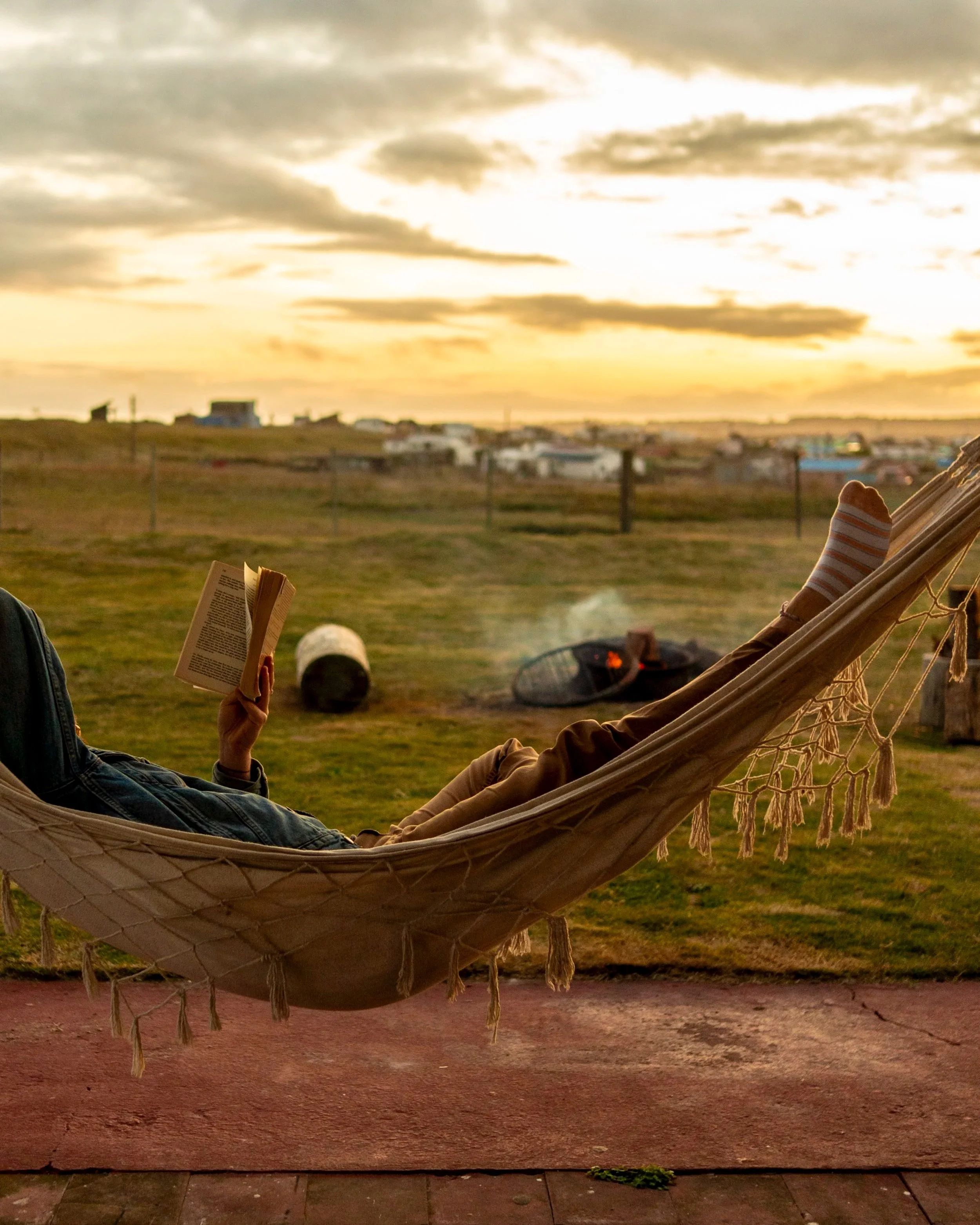 Person lying in a hammock, reading a book, at sunset in a grassy outdoor area with a fire pit and distant houses.