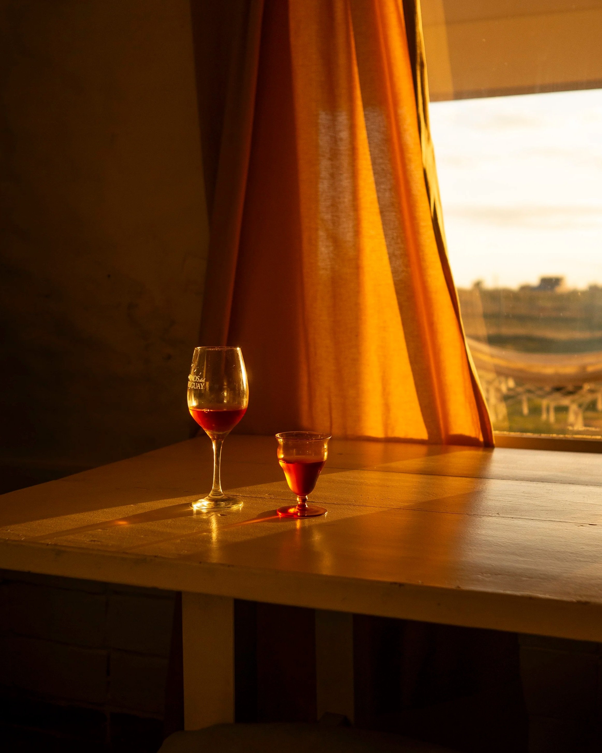 Two glasses filled with red wine on a wooden table near a window with orange curtains, sunlight illuminating the scene.
