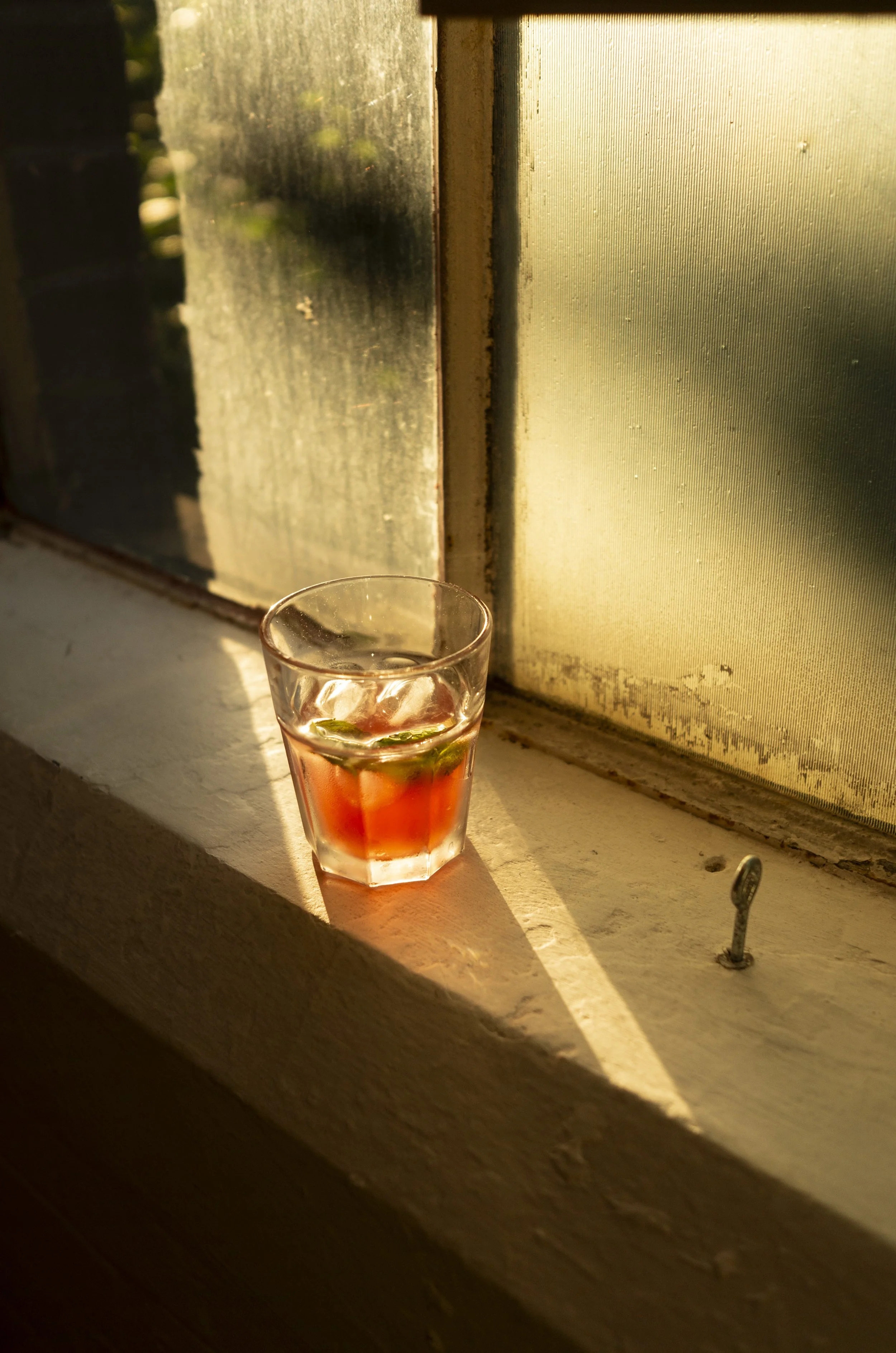 A glass of cold beverage with ice and lime slices on a windowsill bathed in warm sunlight.