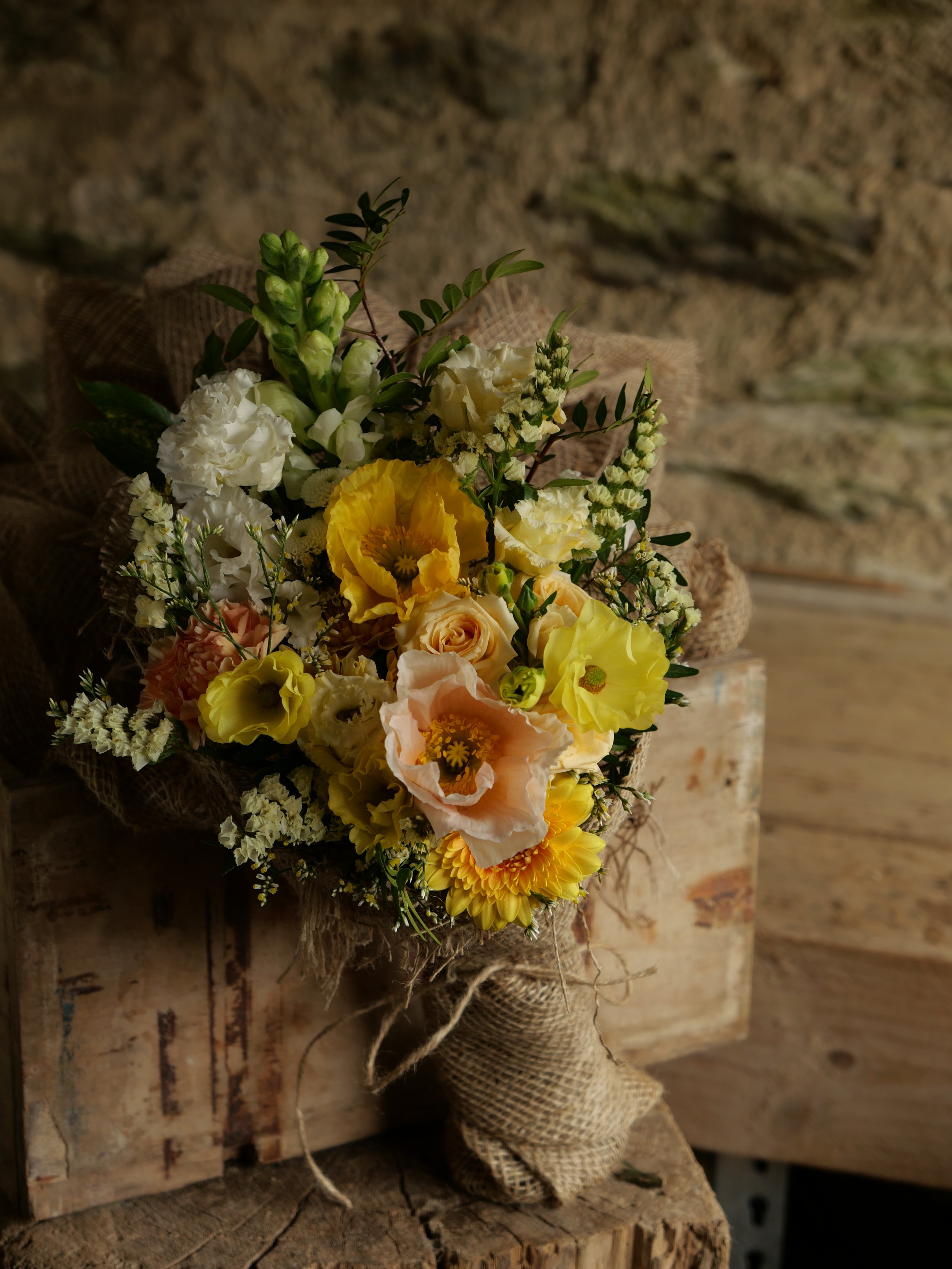 A bouquet of mixed yellow, white, and light peach flowers wrapped in burlap rests on a wooden surface against a rustic stone wall background.