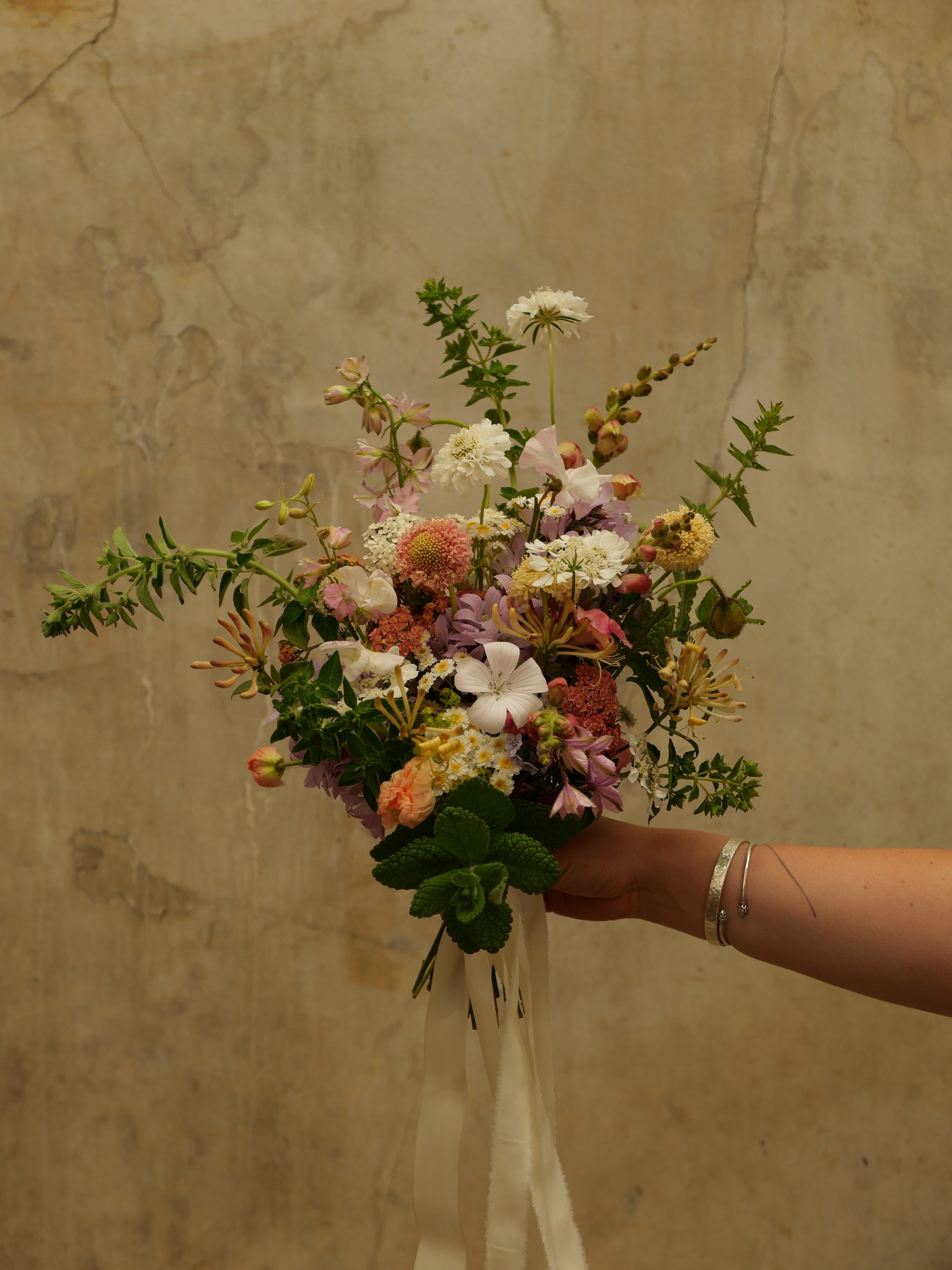A person's hand holding a colorful bouquet of flowers against a textured beige wall.