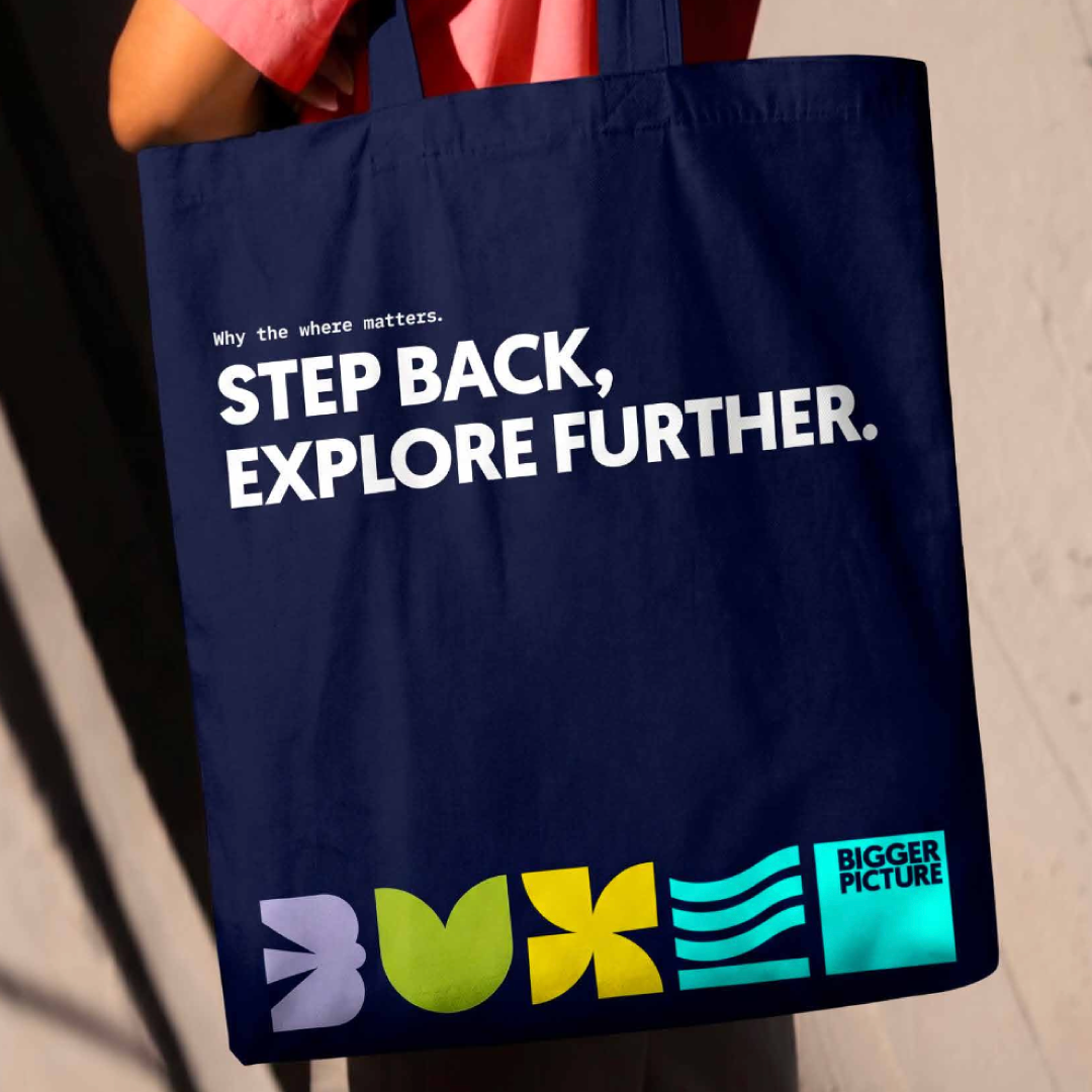 Blue shopping bag with white text that reads "Why the where matters. STEP BACK, EXPLORE FURTHER." and colorful logo design including the words "BIGGER PICTURE".