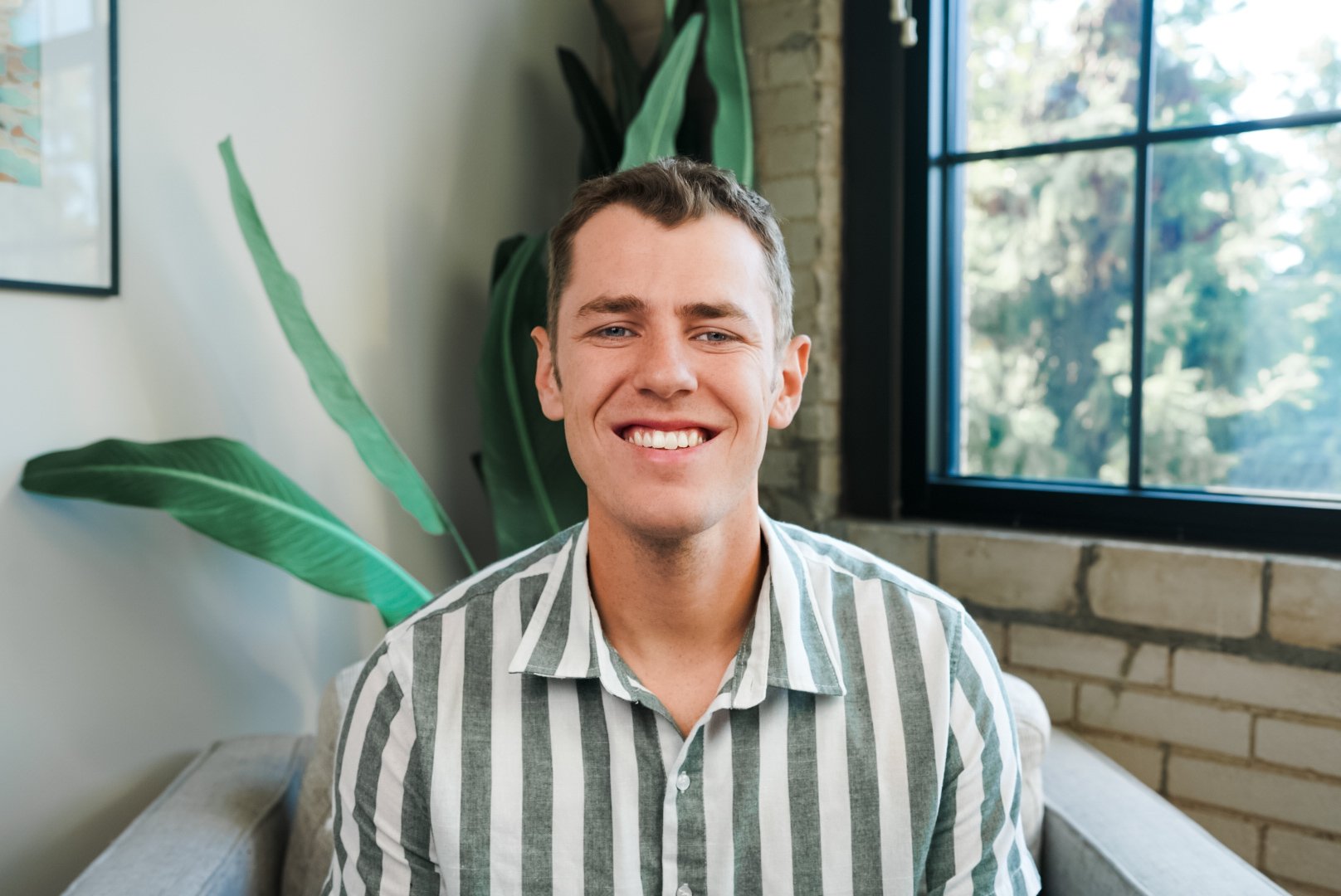 A young man with short brown hair smiling while sitting on a sofa in a room with a large window and green plants.