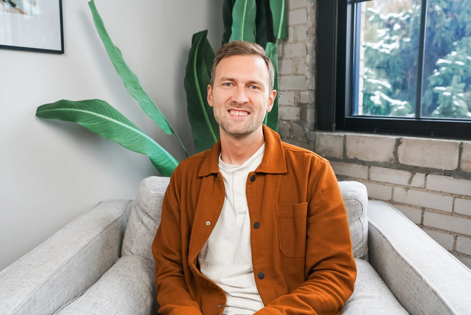 A man sitting on a light gray couch in a modern living room with green plants, brick wall, and a large window showing snow-covered trees outside.