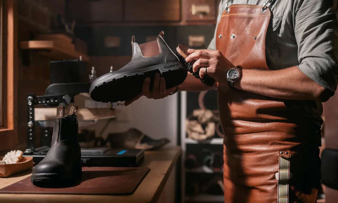 A person in a leather apron is inspecting or cleaning a brown leather boot inside a workshop or shoe store.