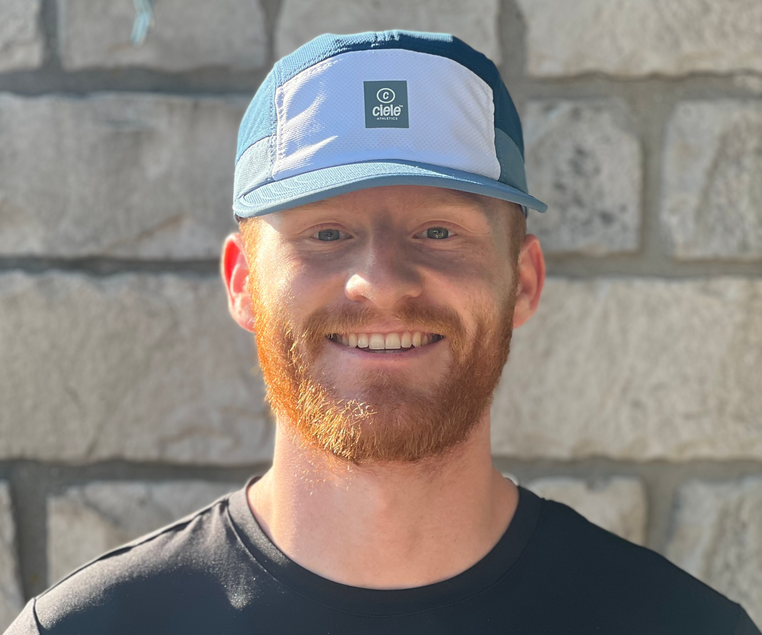 A smiling man with a red beard wearing a black shirt and a blue and white cap, standing outdoors against a stone wall.