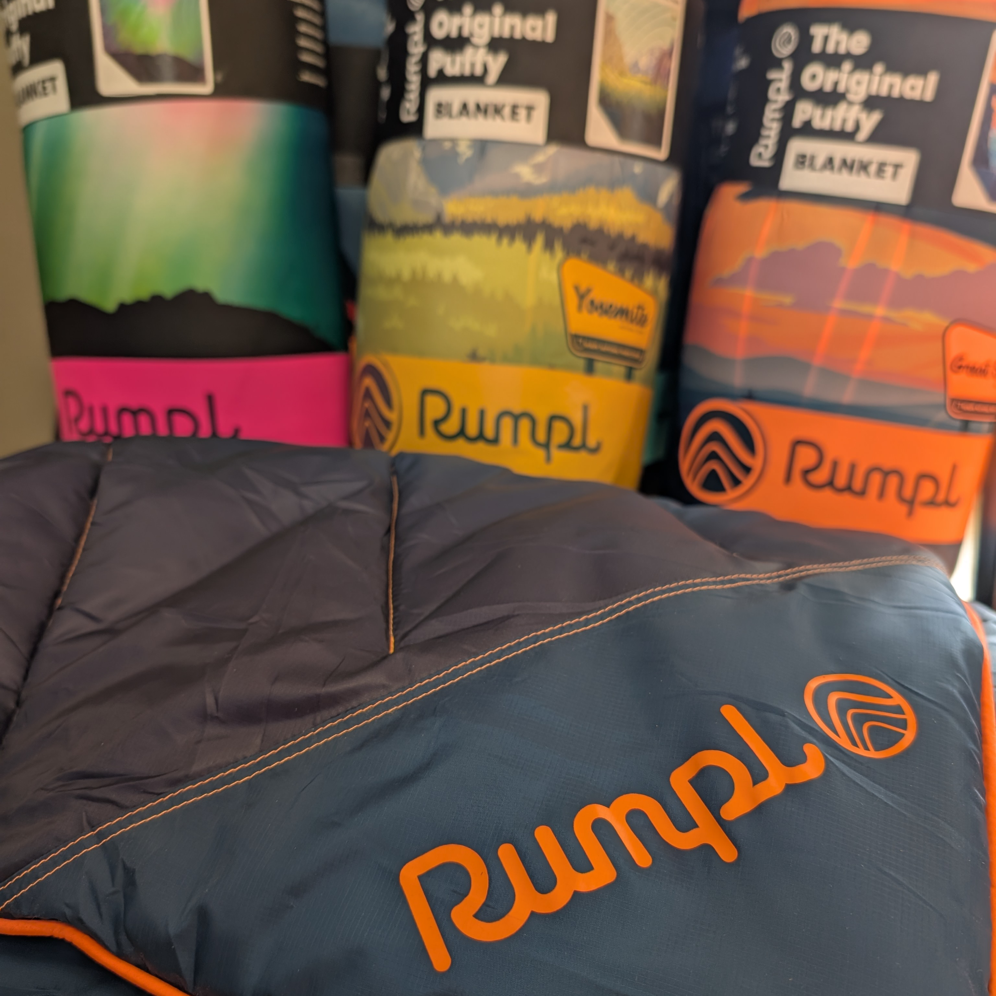 Rumpel brand sleeping bags in various colorful designs behind a dark gray down sleeping bag with orange piping and orange 'Rumpel' logo in the foreground.