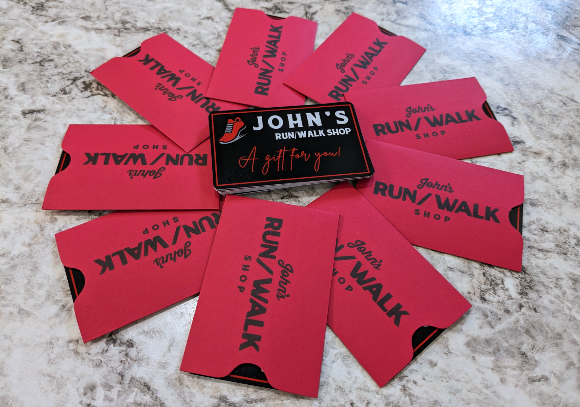 A circular arrangement of red gift cards from John’s Run/Walk Shop with a black box in the center that reads "John’s Run/Walk Shop - A gift for you!" on a marble surface.