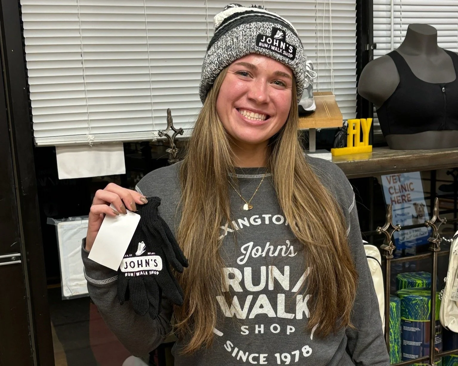 A smiling young woman with long hair wearing a beanie and a gray sweatshirt, holding a pair of black gloves with a logo from John's Run/Walk Shop inside a store with various merchandise.