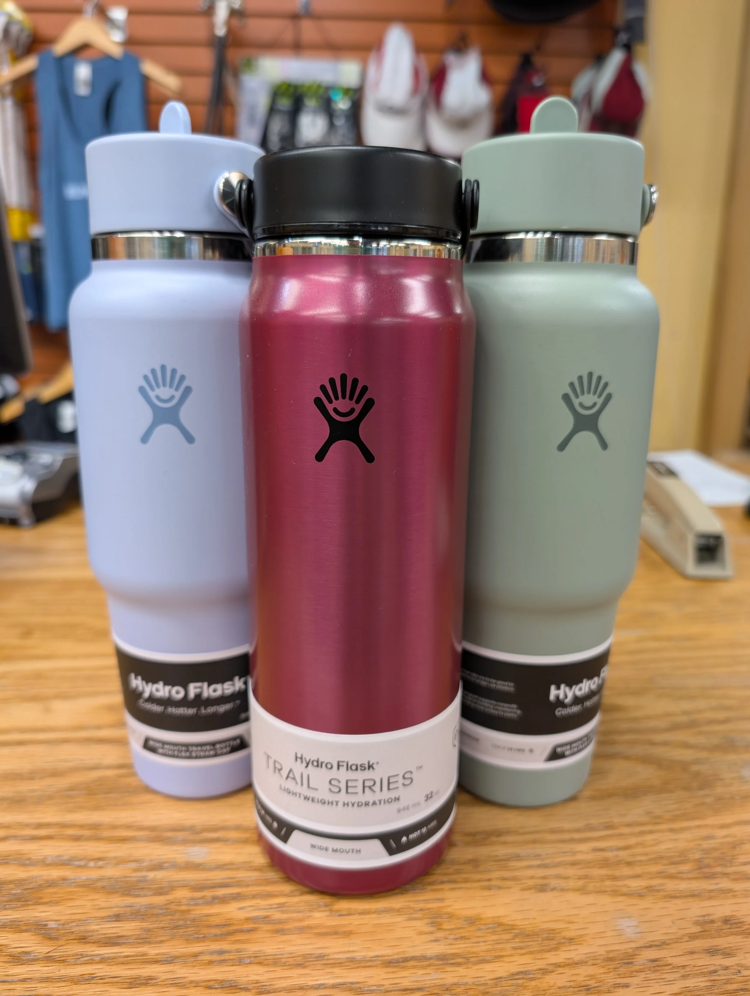 Three Hydro Flask water bottles in light blue, pink, and light green colors standing on a wooden surface in a store display.