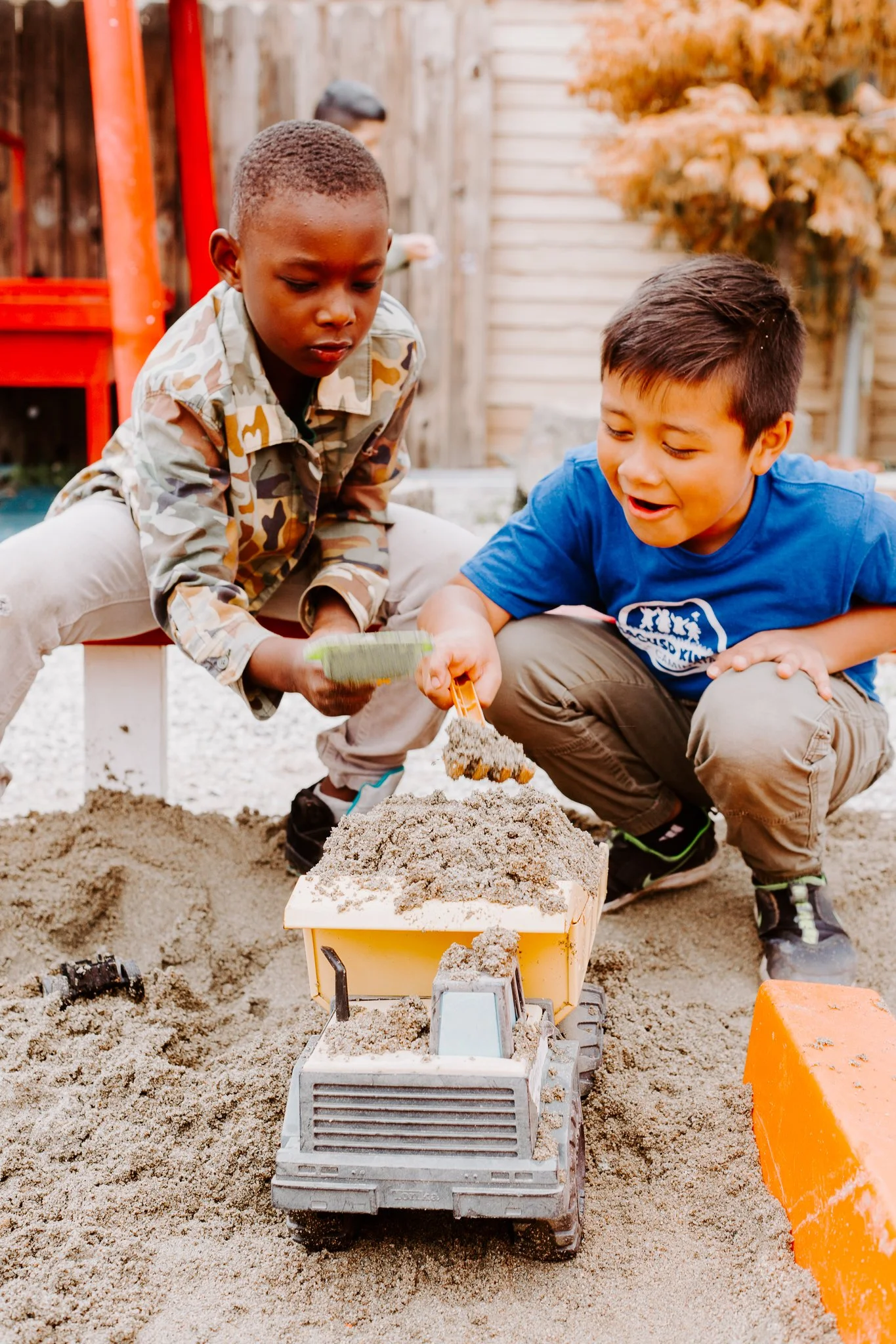 Two boys playing in a sandbox, using toy construction equipment to move sand.
