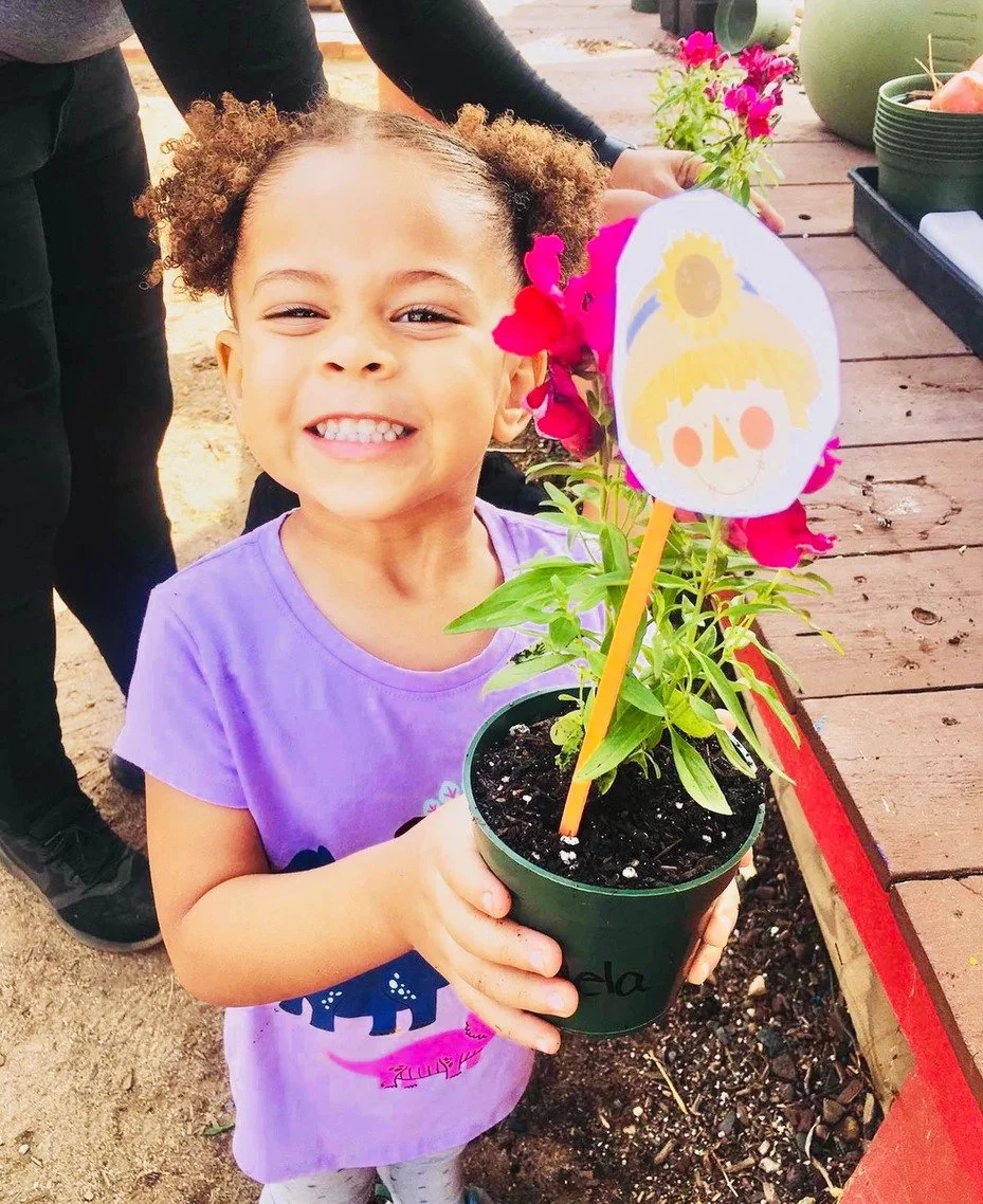 A young girl smiling and holding a pot with a flowering plant and colorful decorations, including a paper sun and smiling face.