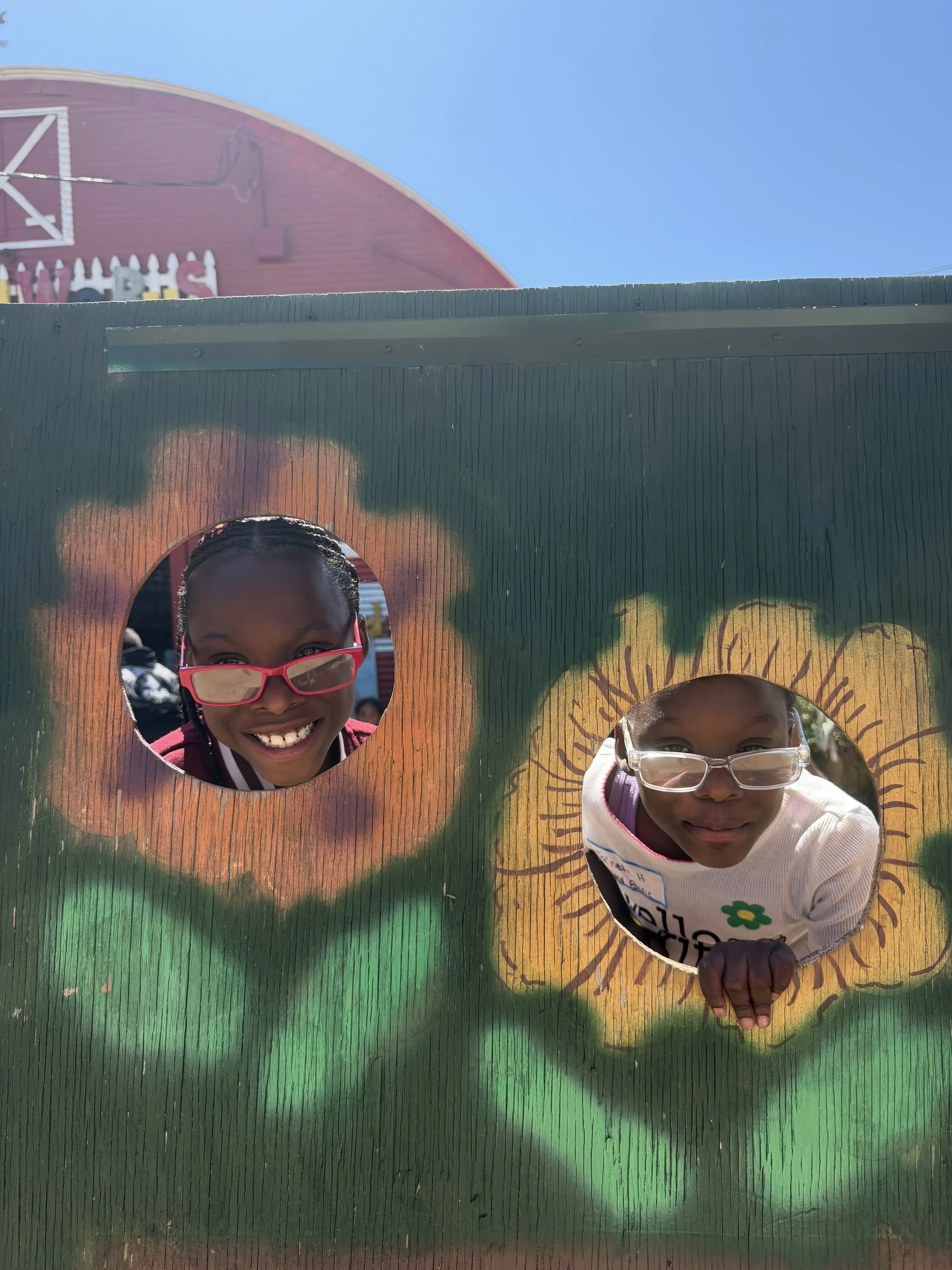 Two children looking through heart-shaped openings in a colorful painted wooden board at an outdoor event, with a red barn and a clear blue sky in the background.