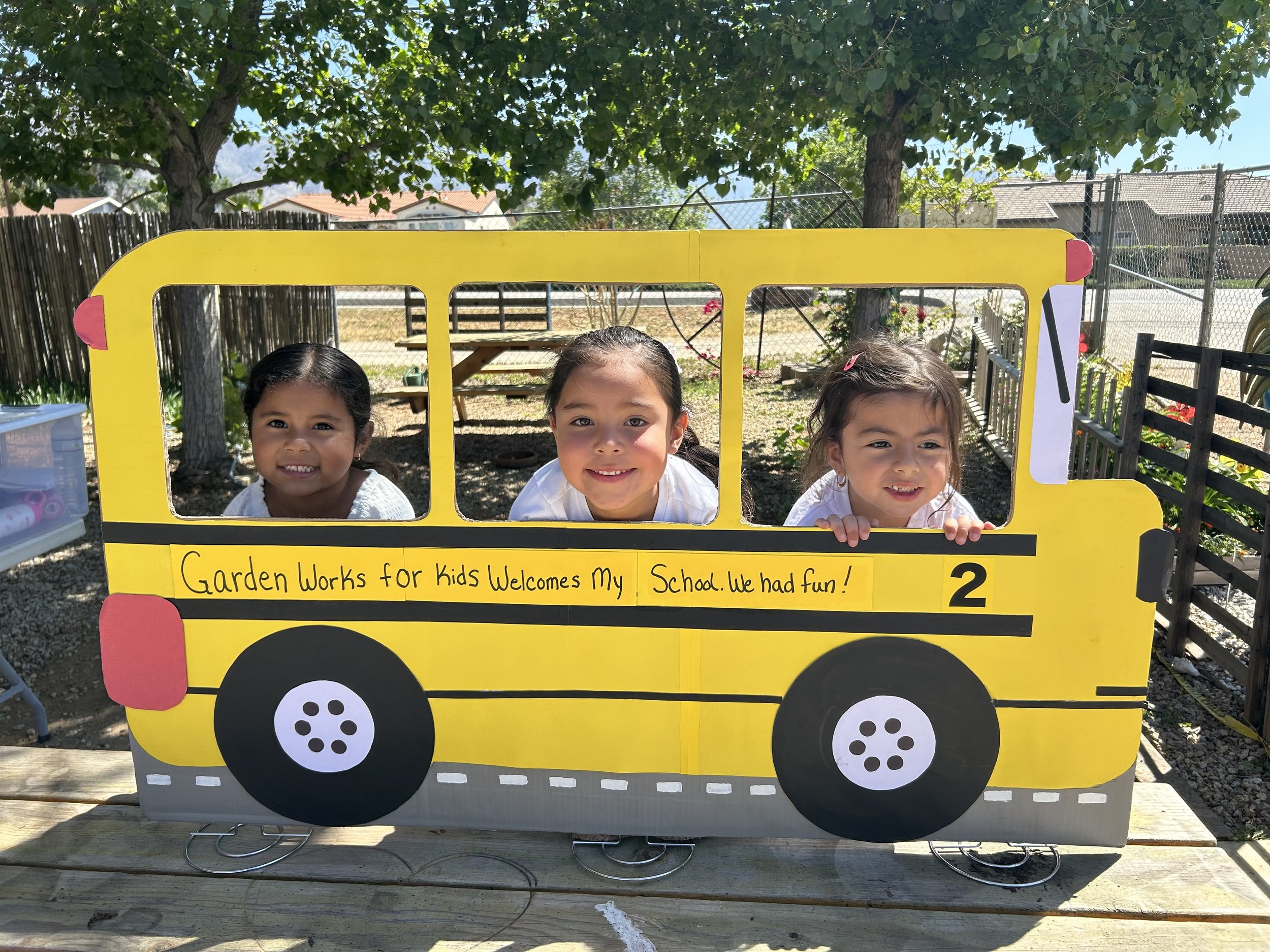 Kids are inside a yellow, bus-shaped photo prop with black wheels. The prop has a note that says "Garden Works for Kids Welcomes My School. We had fun!". They are outdoors, under a tree with a chain-link fence in the background.