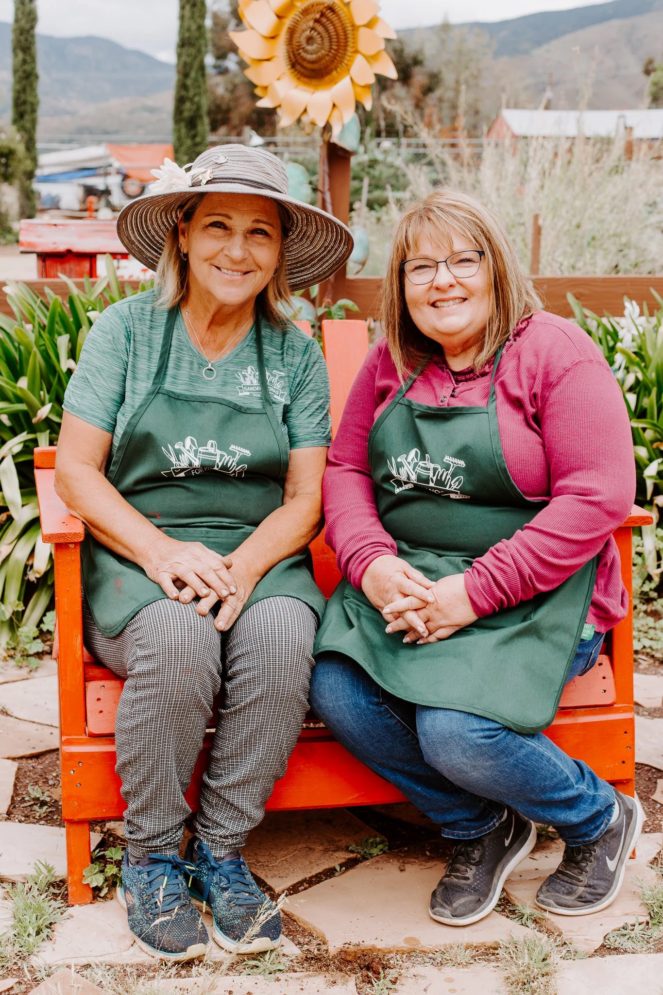 Two women sitting on an orange bench outdoors, wearing green aprons, smiling, with a garden and mountains in the background.