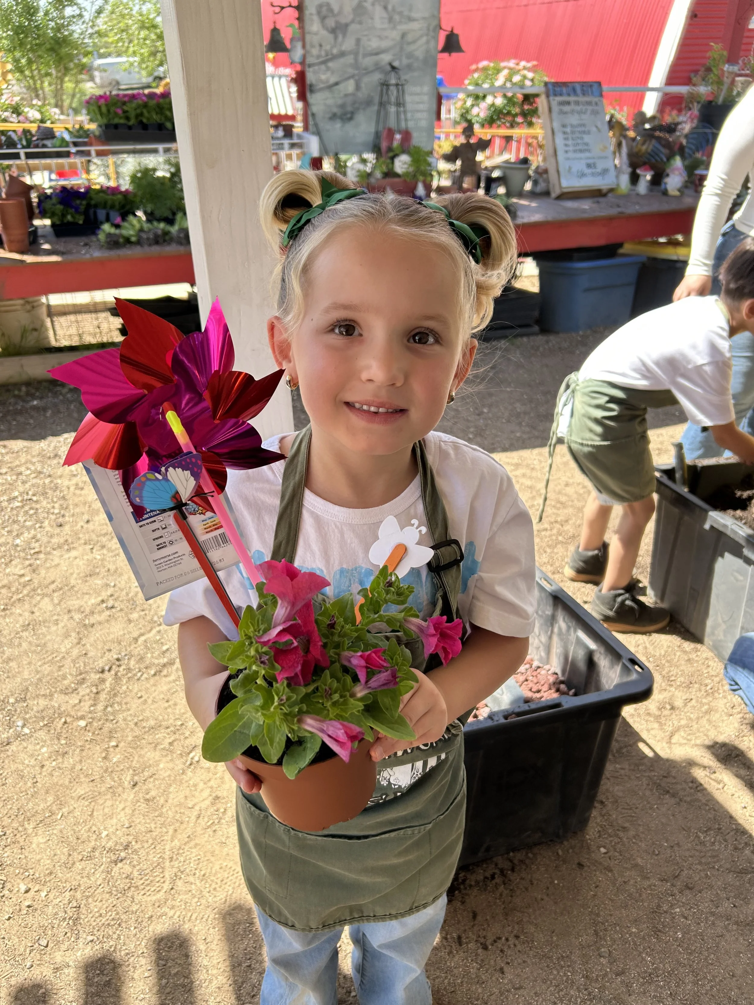 A young girl with blonde hair in two buns, green ribbons, and earrings, holding a potted pink and purple flowering plant at a garden center or nursery. She is smiling and wearing a white shirt, an apron, and green pants. In the background, there are 