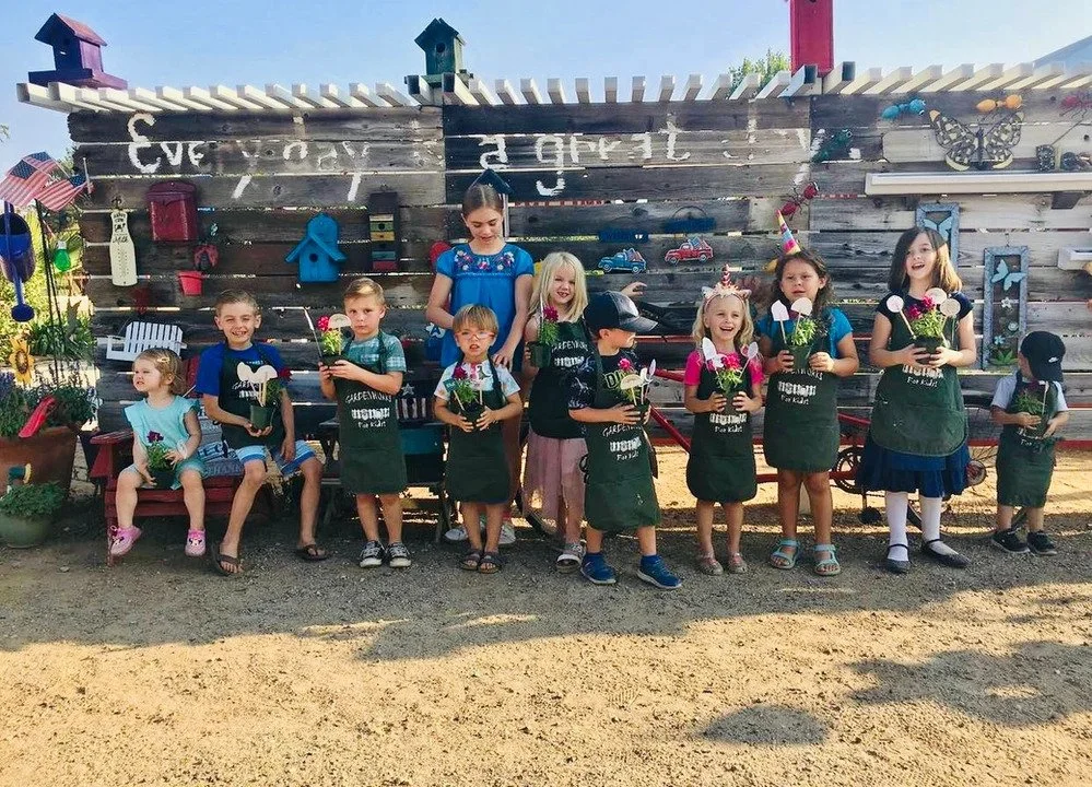 Group of children and a woman standing in front of a wooden wall holding potted plants, with some children sitting on a bench, all smiling. The setting appears to be a garden or outdoor area.