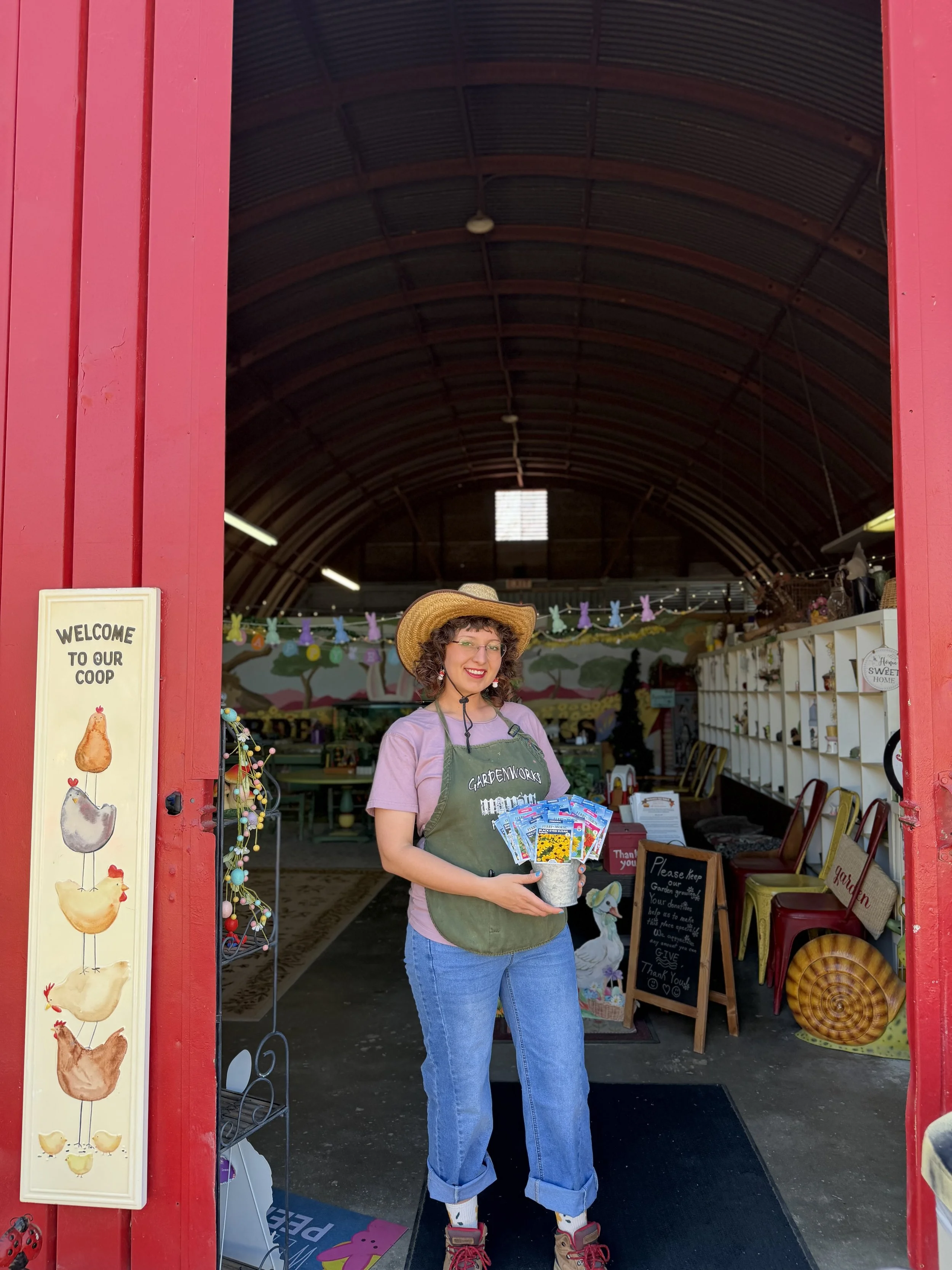 A smiling woman standing in the entrance of a farm shop, holding seed packets. She is wearing a straw hat, glasses, a purple T-shirt, and jeans. Inside the shop, colorful decorations and various items are visible.