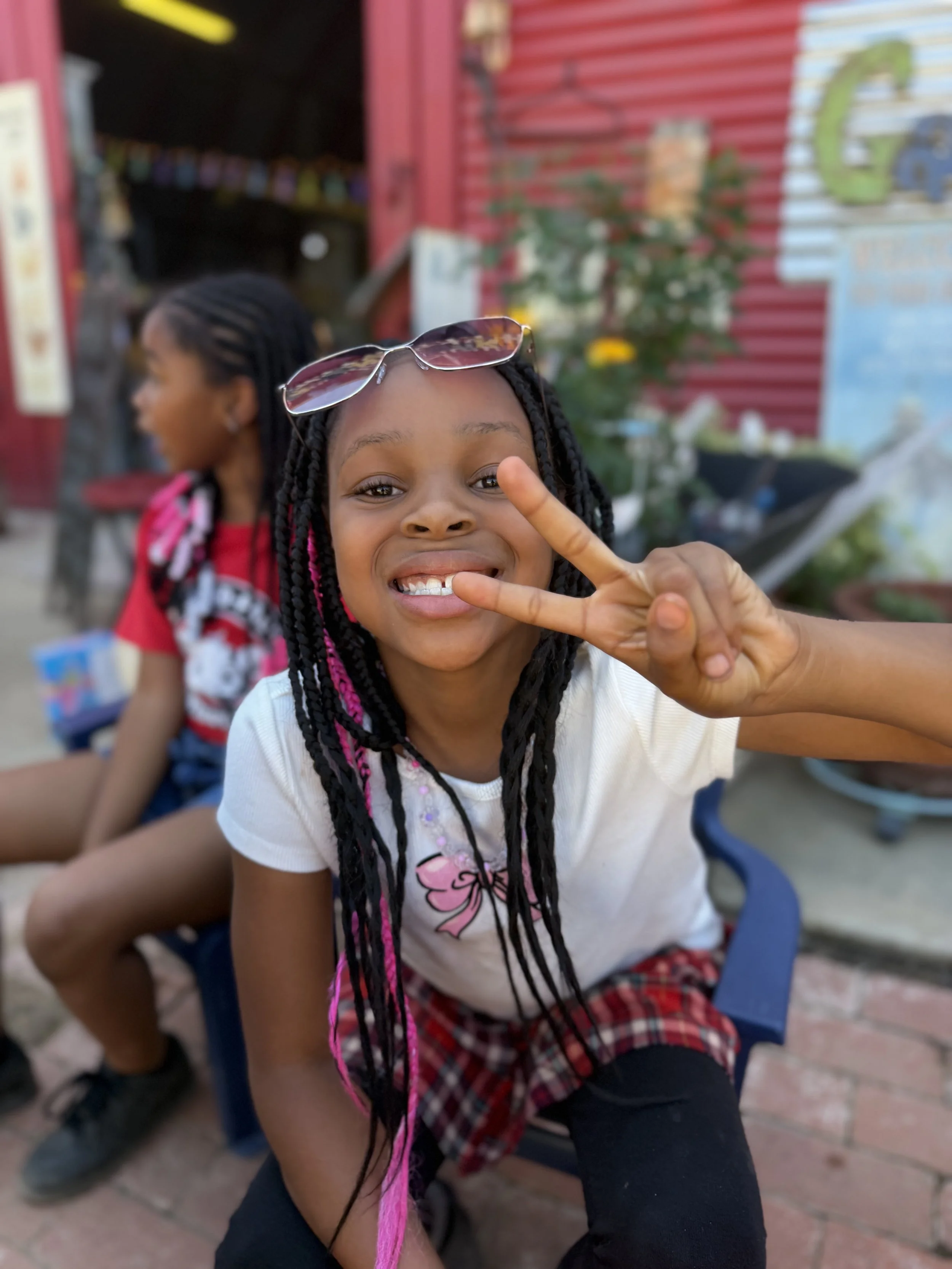 A young girl with braided hair and pink highlights smiling and flashing a peace sign, sitting outside on a blue chair in front of a red and blue building with plants and graffiti.