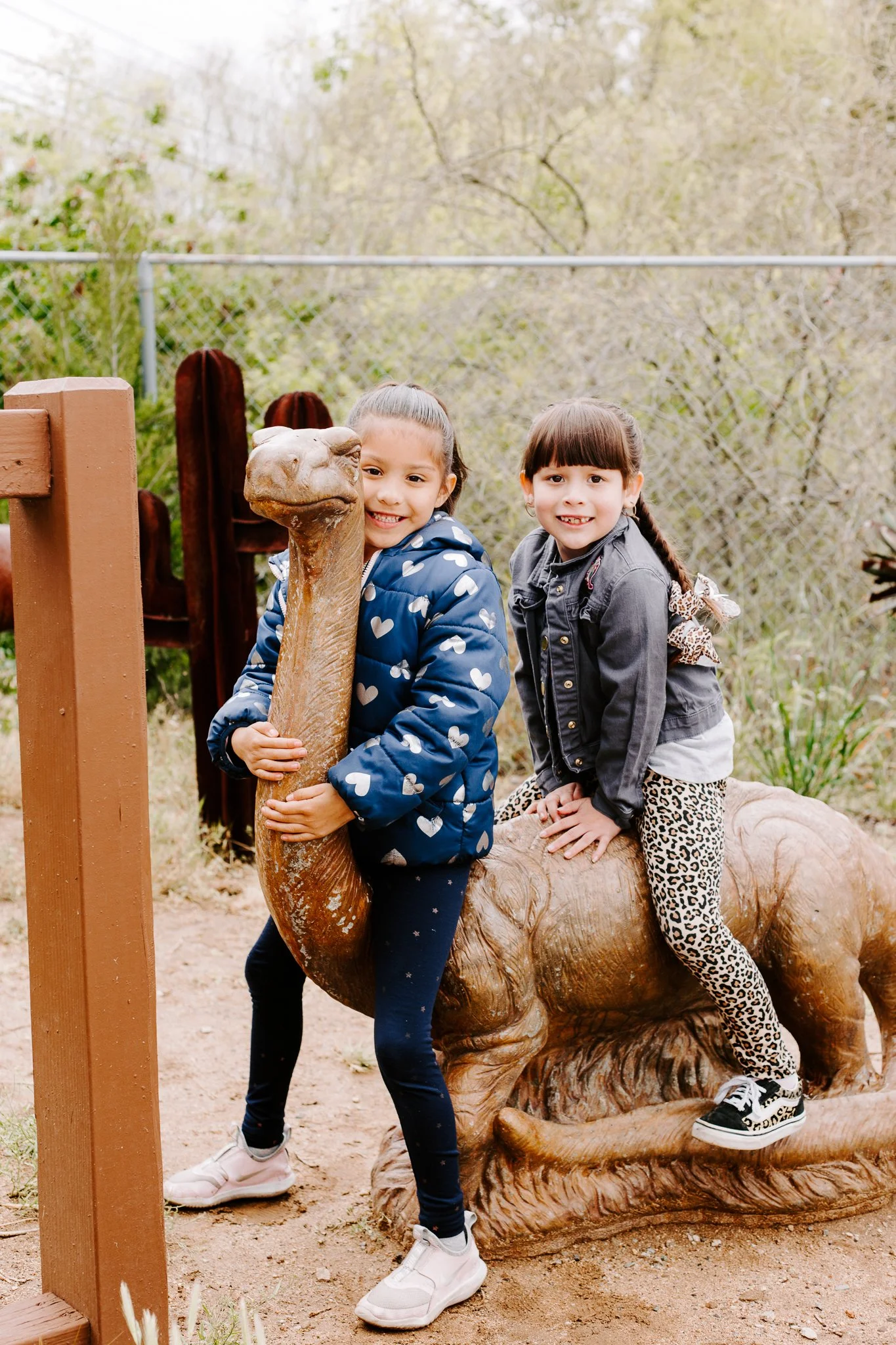 Two young girls riding a wooden dinosaur sculpture at an outdoor park.