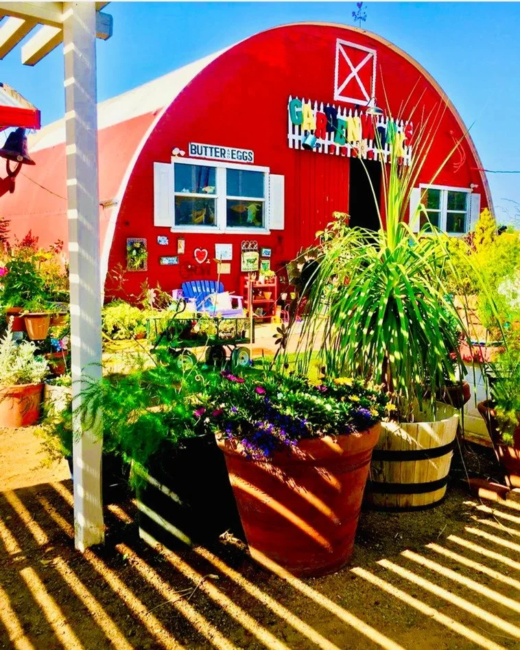 Red barn with colorful garden and potted plants in front, decorated with signs including 'Butter Eggs' and a sign with the word 'GARDEN' in multicolored letters.