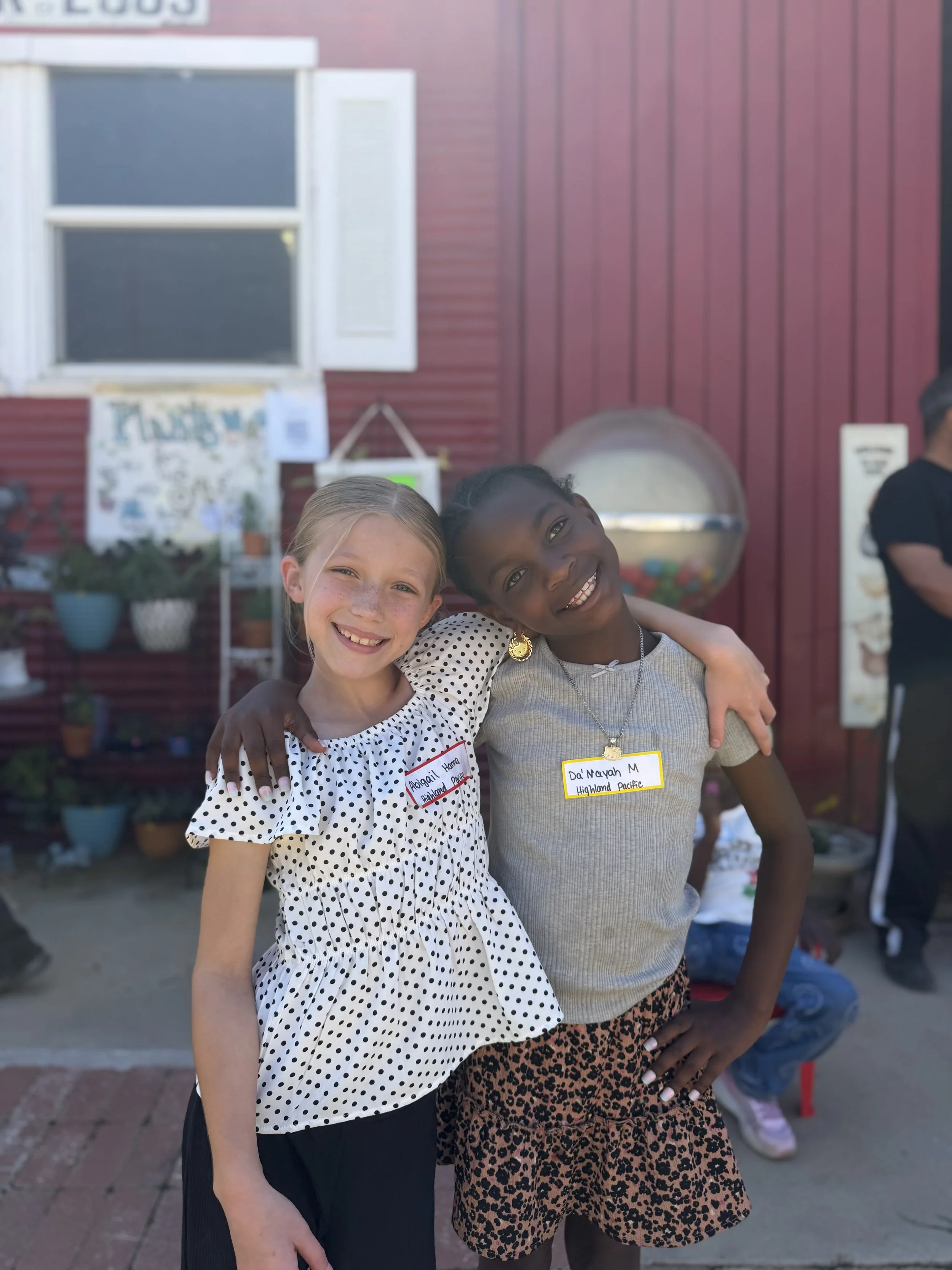 Two young girls smiling with arms around each other, standing outdoors in front of a red building with potted plants, wearing casual clothing and name tags.