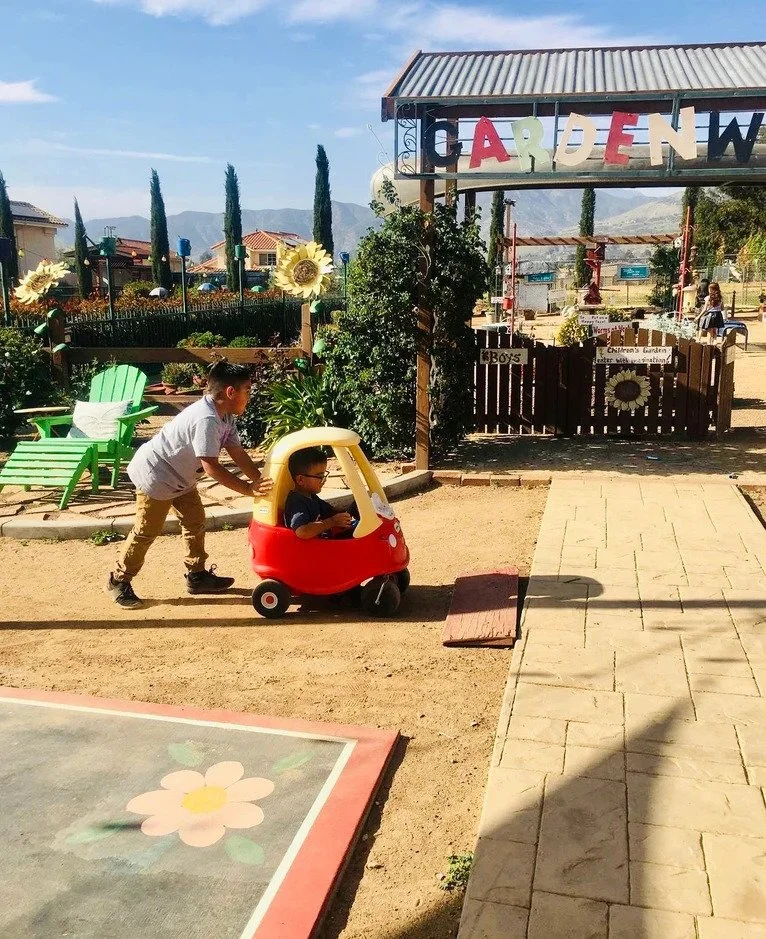 Two boys are outdoors in a playground. One boy is pushing a small red and yellow toy car with a child sitting inside. The playground has green chairs, flowers, trees, and a sign reading 'GARDEN' above a fence. There is a building and mountains in the